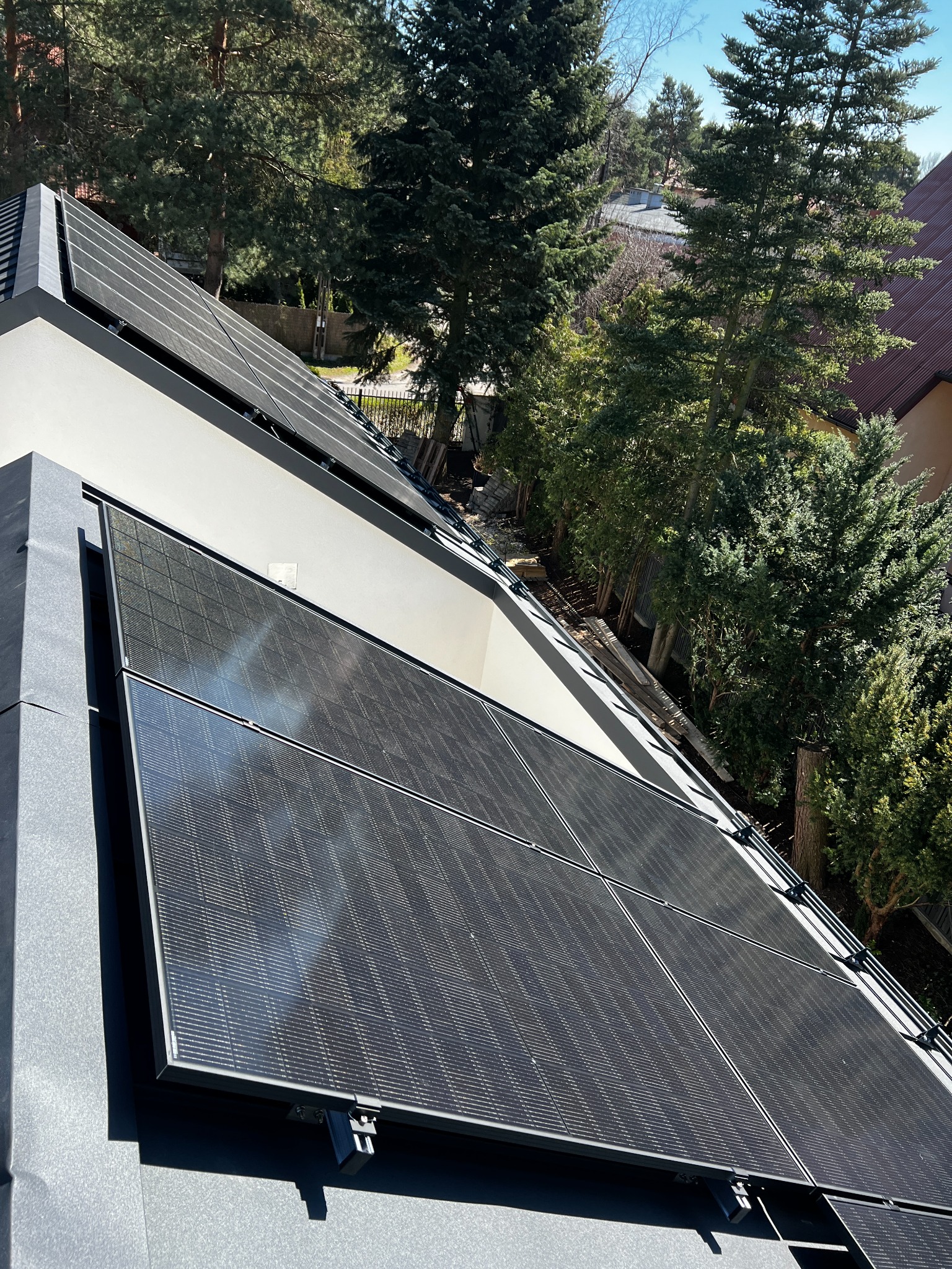 Panele fotowoltaiczne na dachu nowoczesnego domu, widok z góry, otoczenie drzew, słoneczny dzień. Czarny kolor paneli kontrastuje z jasną elewacją budynku.