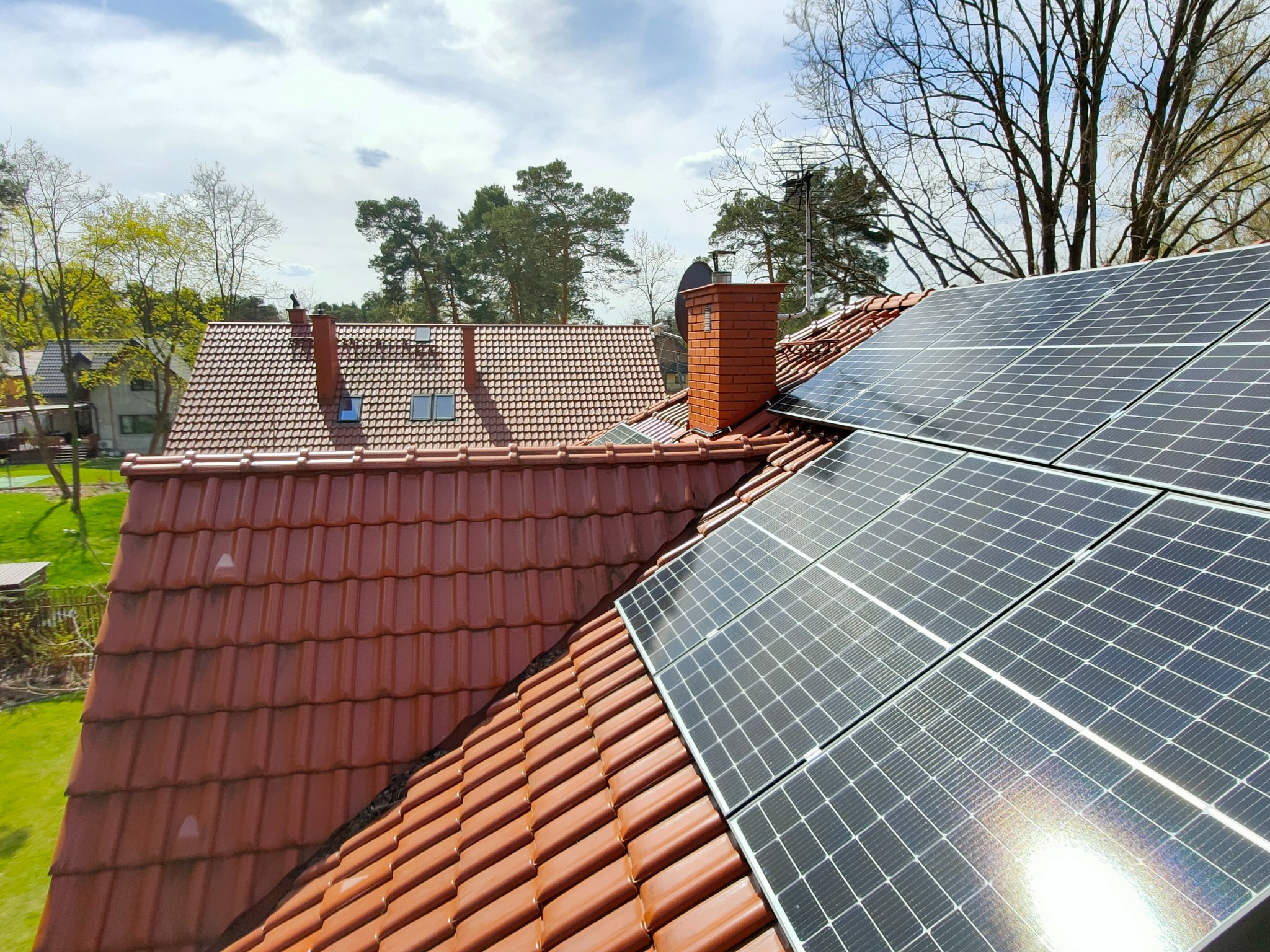 Panele słoneczne na dachu z czerwonej dachówki, widok z góry, w tle budynki i drzewa. Instalacja fotowoltaiczna na tle błękitnego nieba z chmurami.