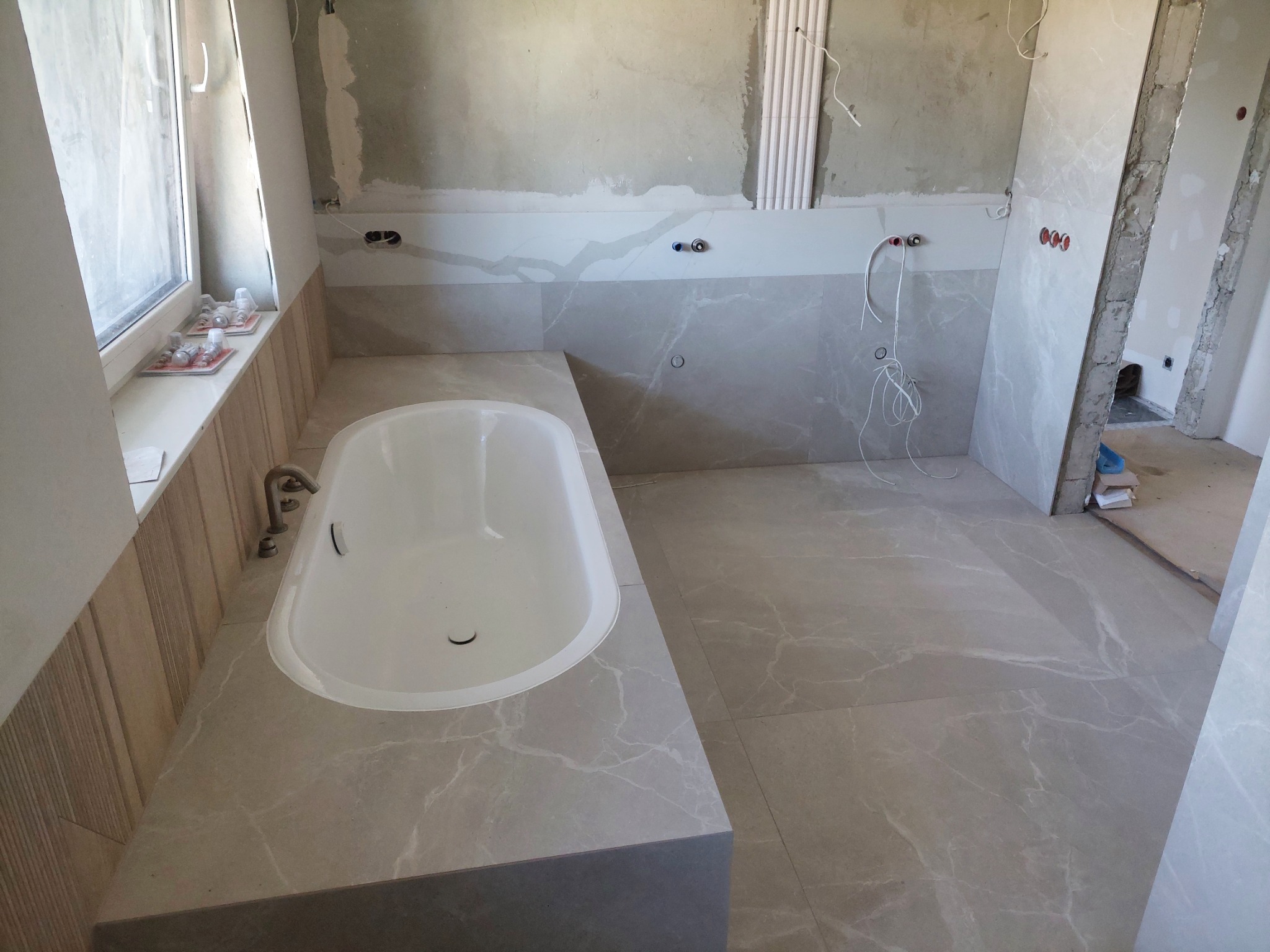 Łazienka w trakcie wykończenia: biała wanna osadzona w obudowie z płytek imitujących marmur, na podłodze i ścianach płytki w podobnym stylu, widoczne rury i przewody elektryczne, okno z akcesoriami...