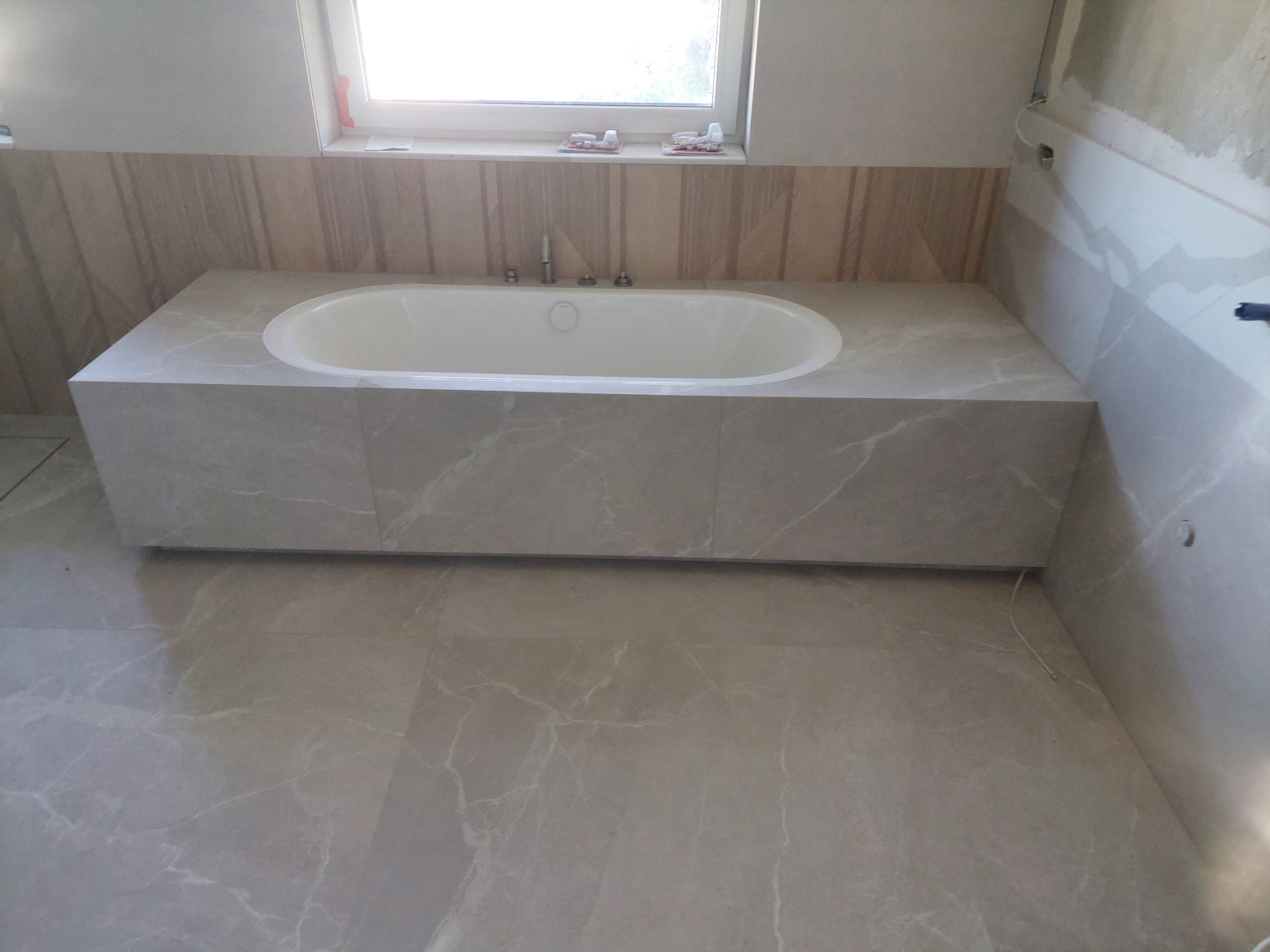 Gotowa obudowa wanny z szarych płyt marmuropodobnych, dopasowana do podłogi i części ścian, widoczne baterie na wannie i okno z akcesoriami na parapecie.