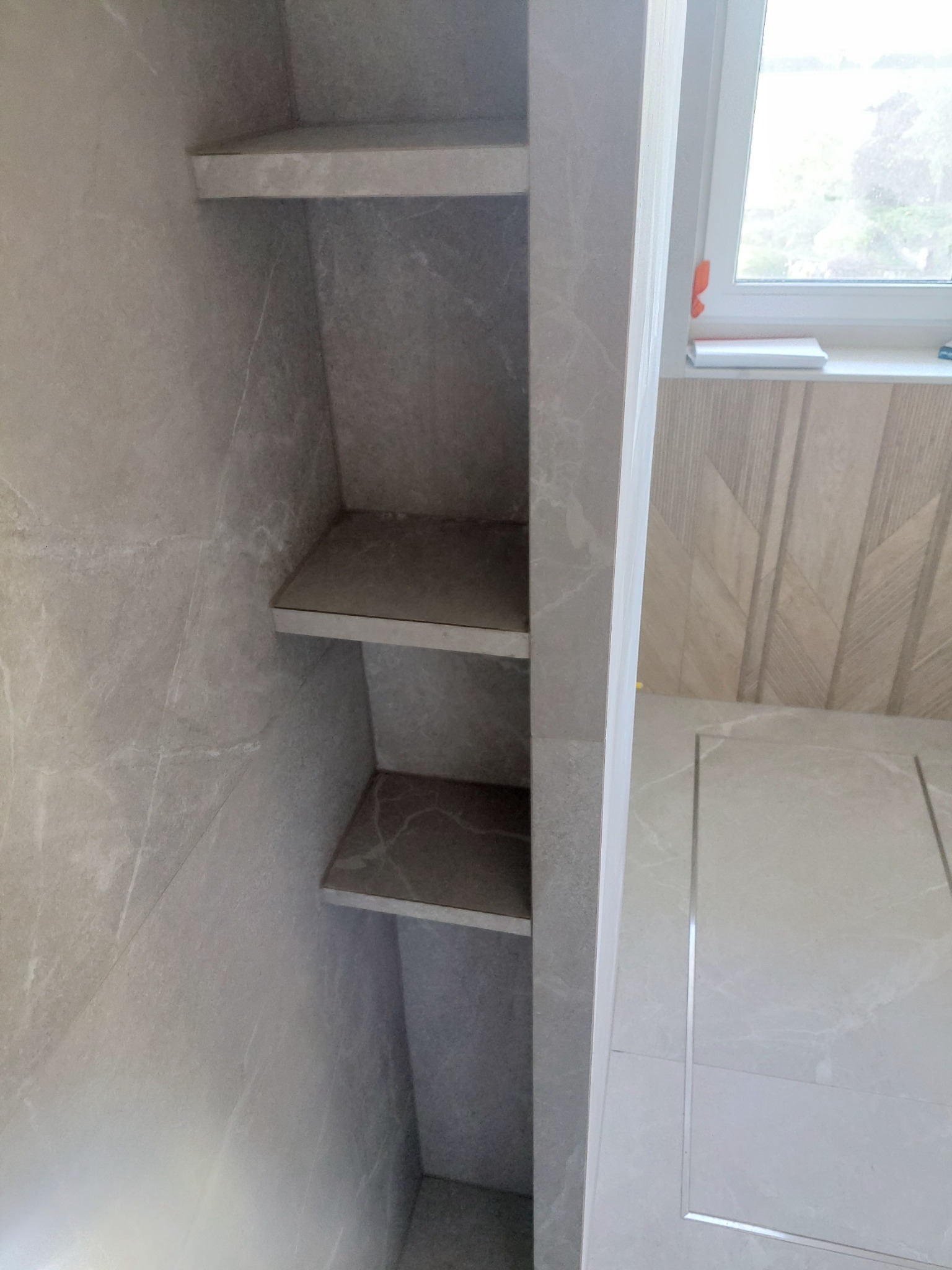 Wnęka z półkami wyłożona szarymi płytkami imitującymi marmur, w łazience z jasną, drewnopodobną okładziną ściany i widocznym oknem.