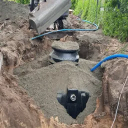 Montaż przydomowej oczyszczalni ścieków w wykopie. Mini koparka zasypuje zbiornik żwirem. Widoczne niebieskie węże i fragment ogrodzenia panelowego w tle.