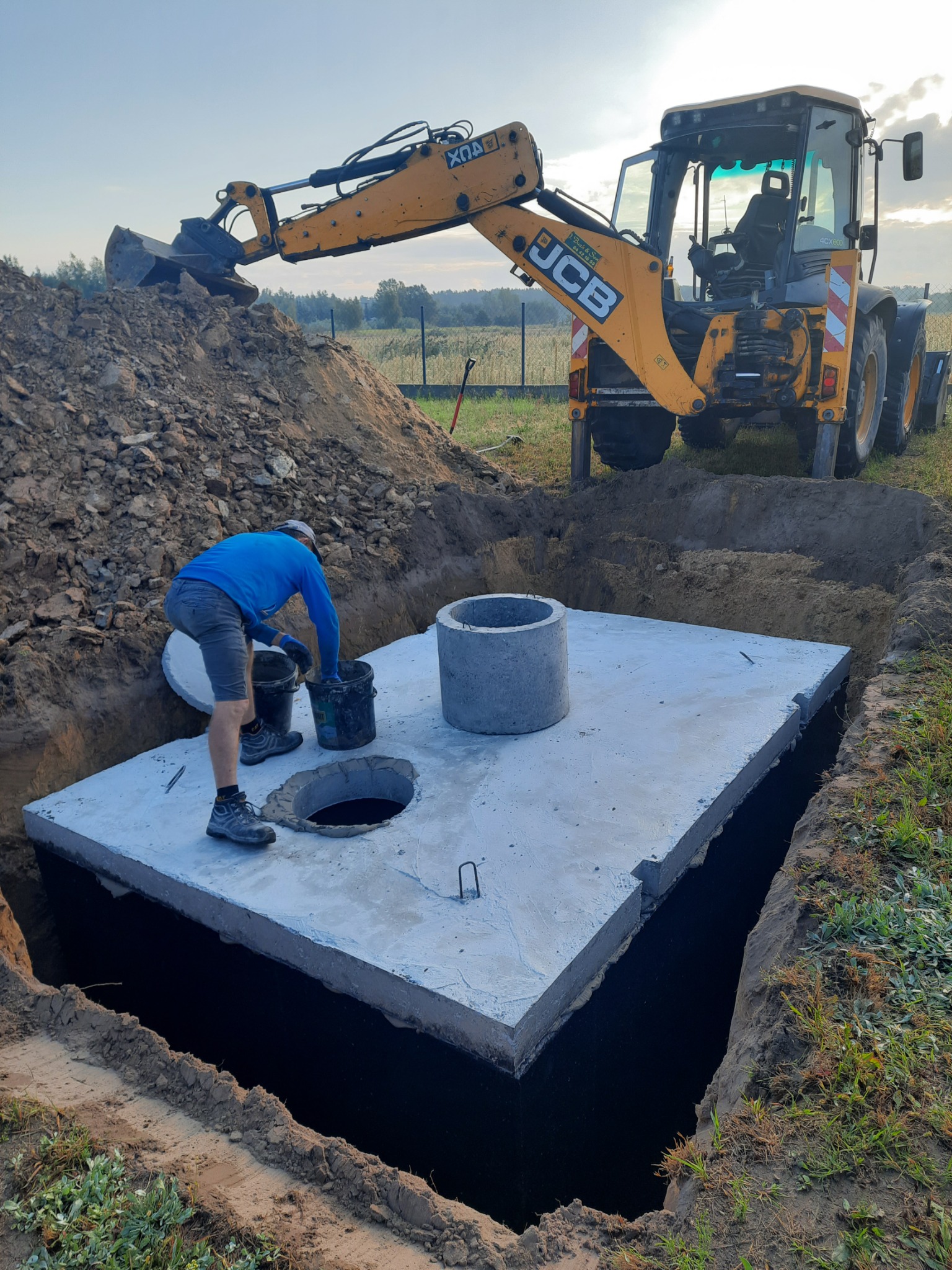 Montaż betonowego szamba: pracownik z wiadrami, wykop, płyta betonowa z otworem, krąg betonowy i koparka JCB w tle.
