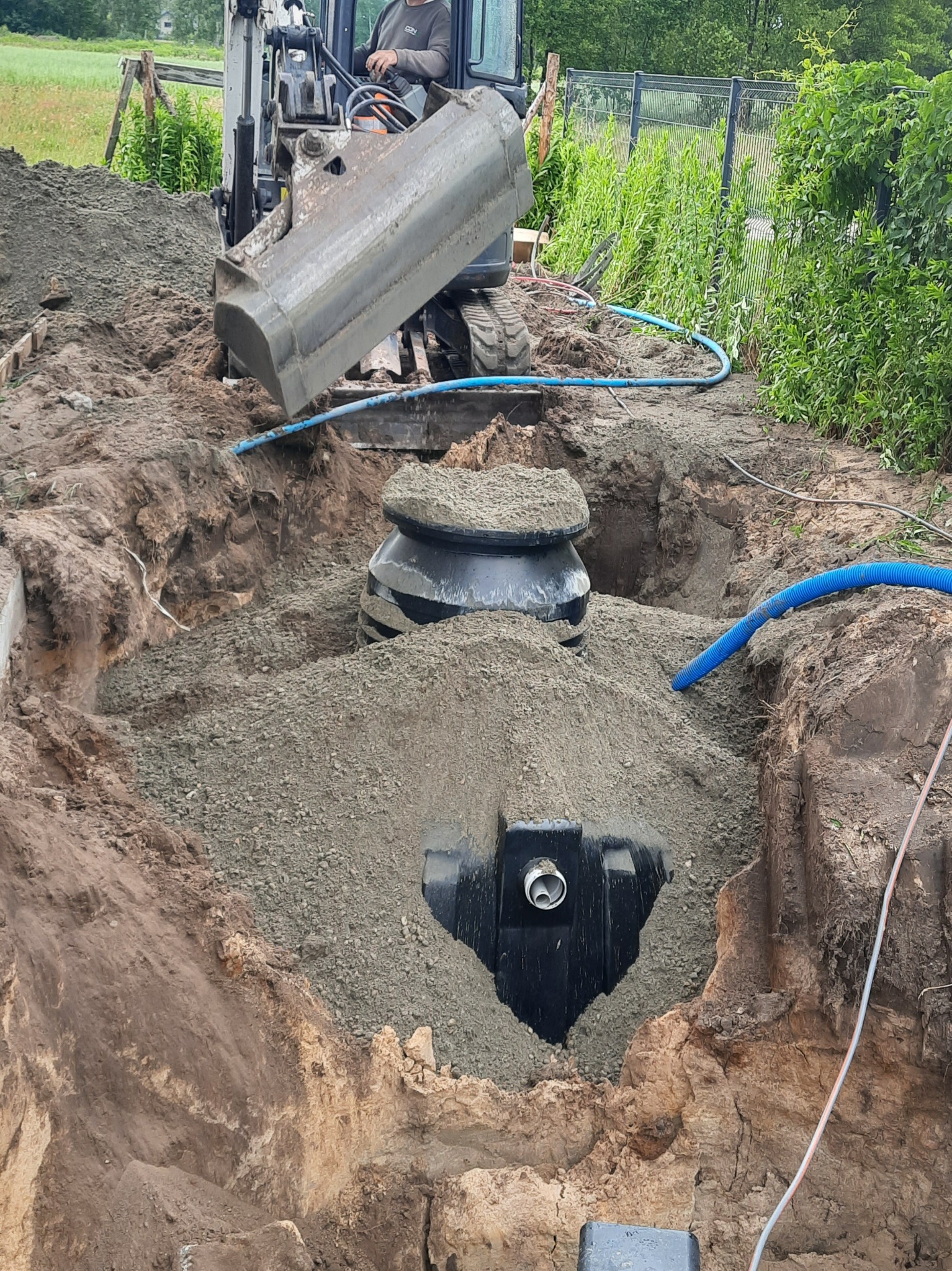 Montaż przydomowej oczyszczalni ścieków w wykopie. Mini koparka zasypuje zbiornik żwirem. Widoczne niebieskie węże i fragment ogrodzenia panelowego w tle.