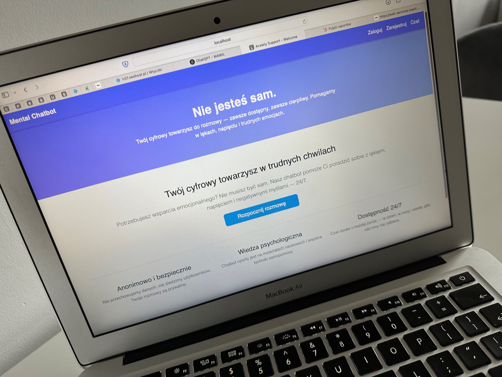 Ekran laptopa MacBook Air wyświetlający stronę internetową Mental Chatbot z hasłem 'Nie jesteś sam' i przyciskiem 'Rozpocznij rozmowę', podkreślającą anonimowość i dostępność 24/7, z widoczną...