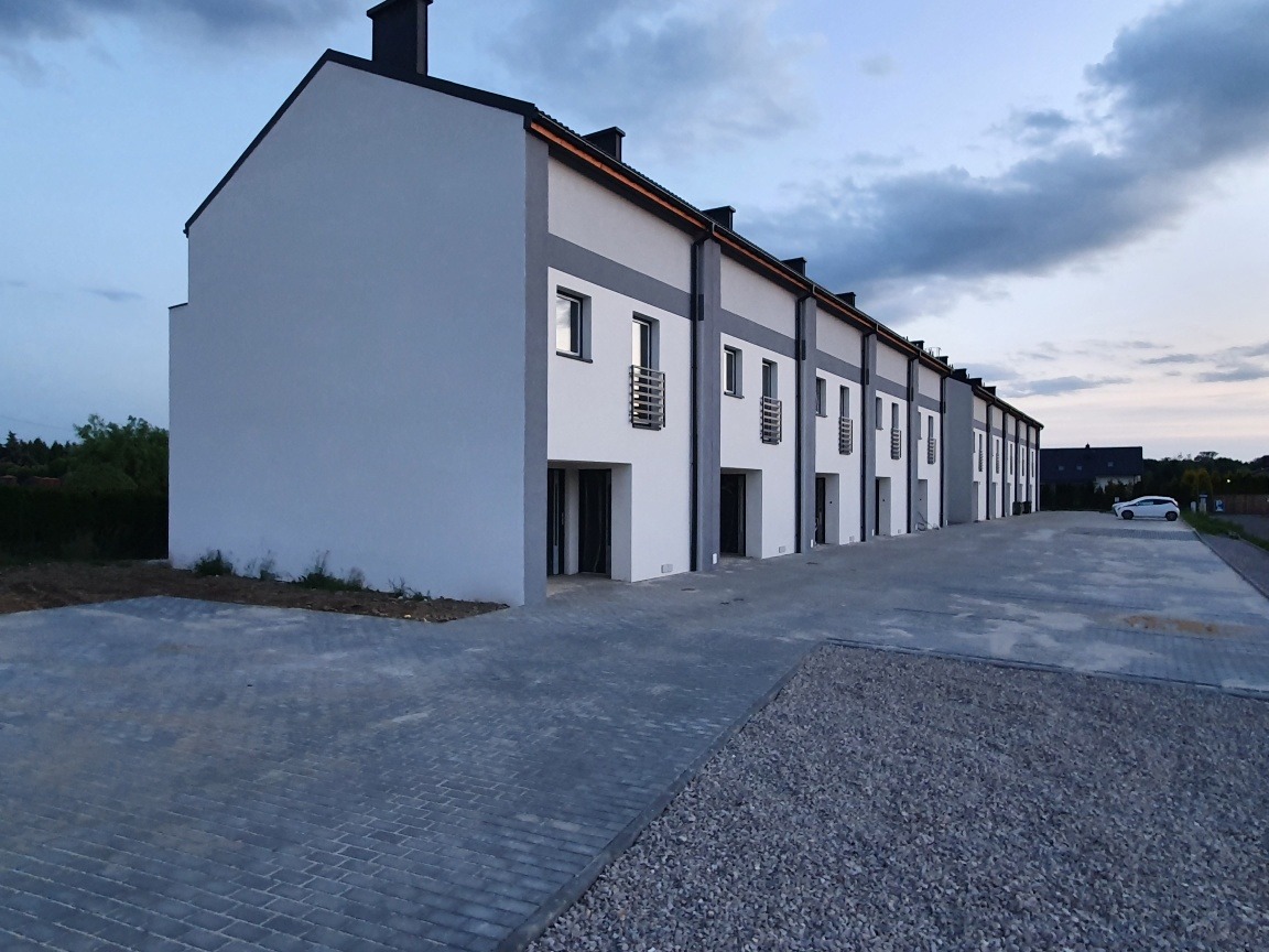 Szereg identycznych, dwukondygnacyjnych domów w minimalistycznym stylu z płaskimi dachami i szarą elewacją, widok z zewnątrz na parking z kostki brukowej i żwiru, w tle zachodzące słońce.