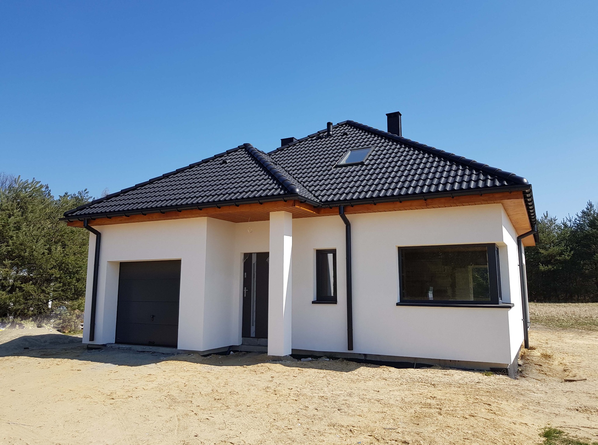 Nowoczesny, parterowy dom jednorodzinny z białymi ścianami, czarną dachówką i grafitową stolarką okienną, usytuowany na działce z piaskiem i widocznym lasem w tle.