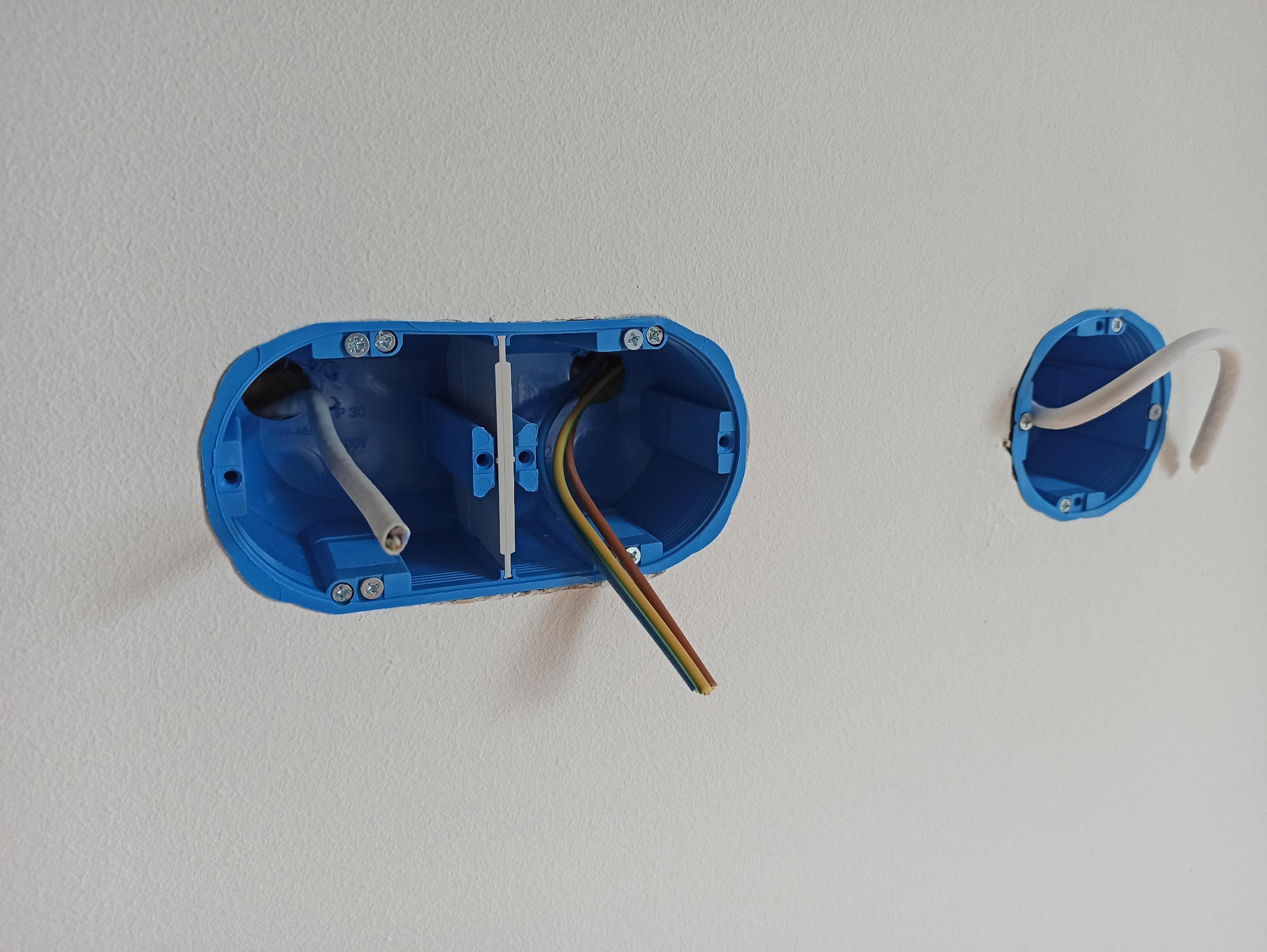 Dwie niebieskie puszki elektryczne zamontowane w białej ścianie, z widocznymi przewodami elektrycznymi wewnątrz.