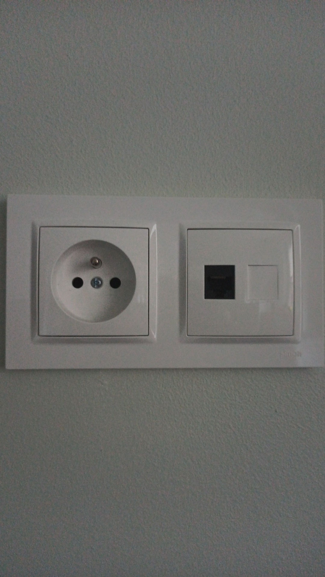 Białe gniazdko elektryczne i gniazdo sieciowe RJ45 zamontowane na szarej ścianie, widoczne delikatne cienie.