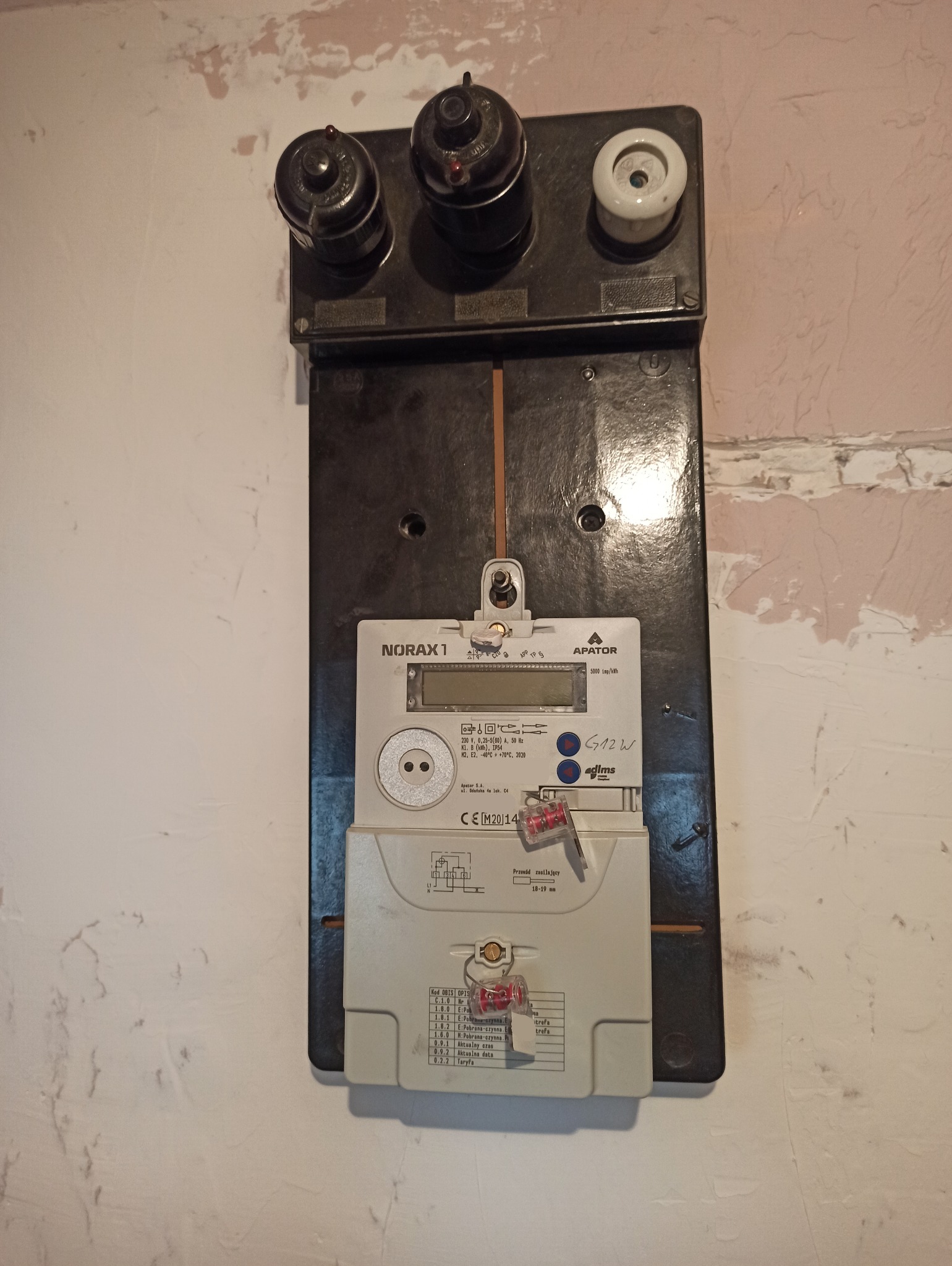 Stara tablica licznikowa z bezpiecznikami topikowymi, zamontowana na ścianie o nierównej fakturze, widoczne ślady ingerencji w instalację.