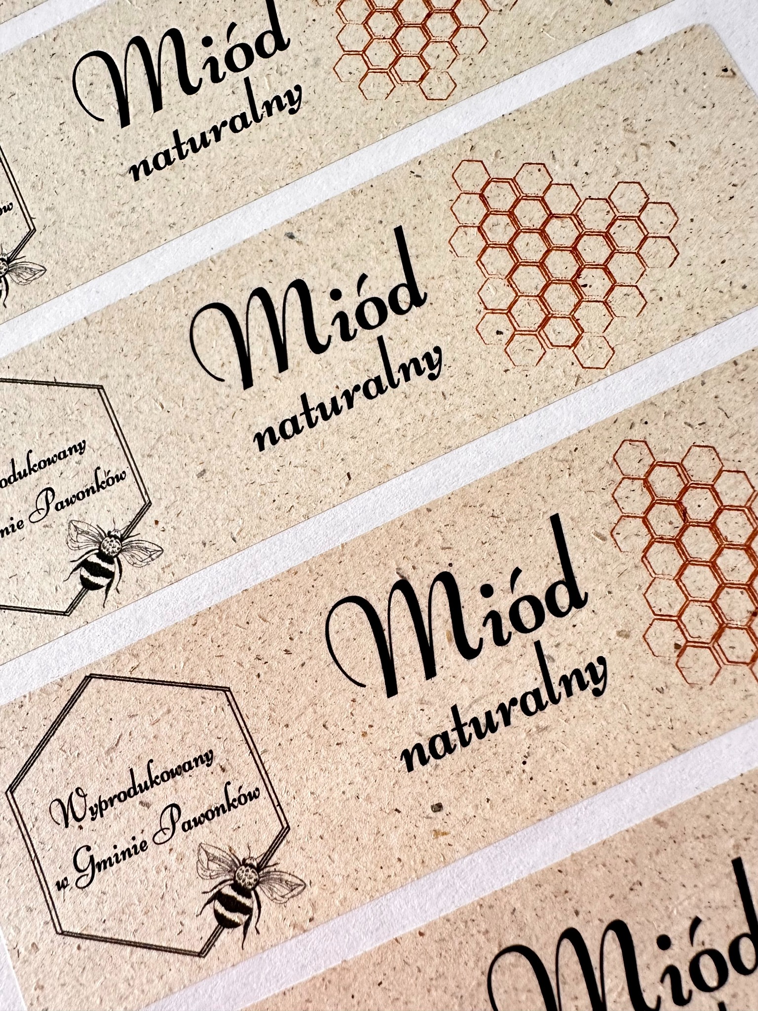 Zbliżenie na rzemieślnicze etykiety na słoiki z miodem naturalnym, z motywem plastra miodu i ilustracją pszczoły, wydrukowane na papierze o fakturze kraft.