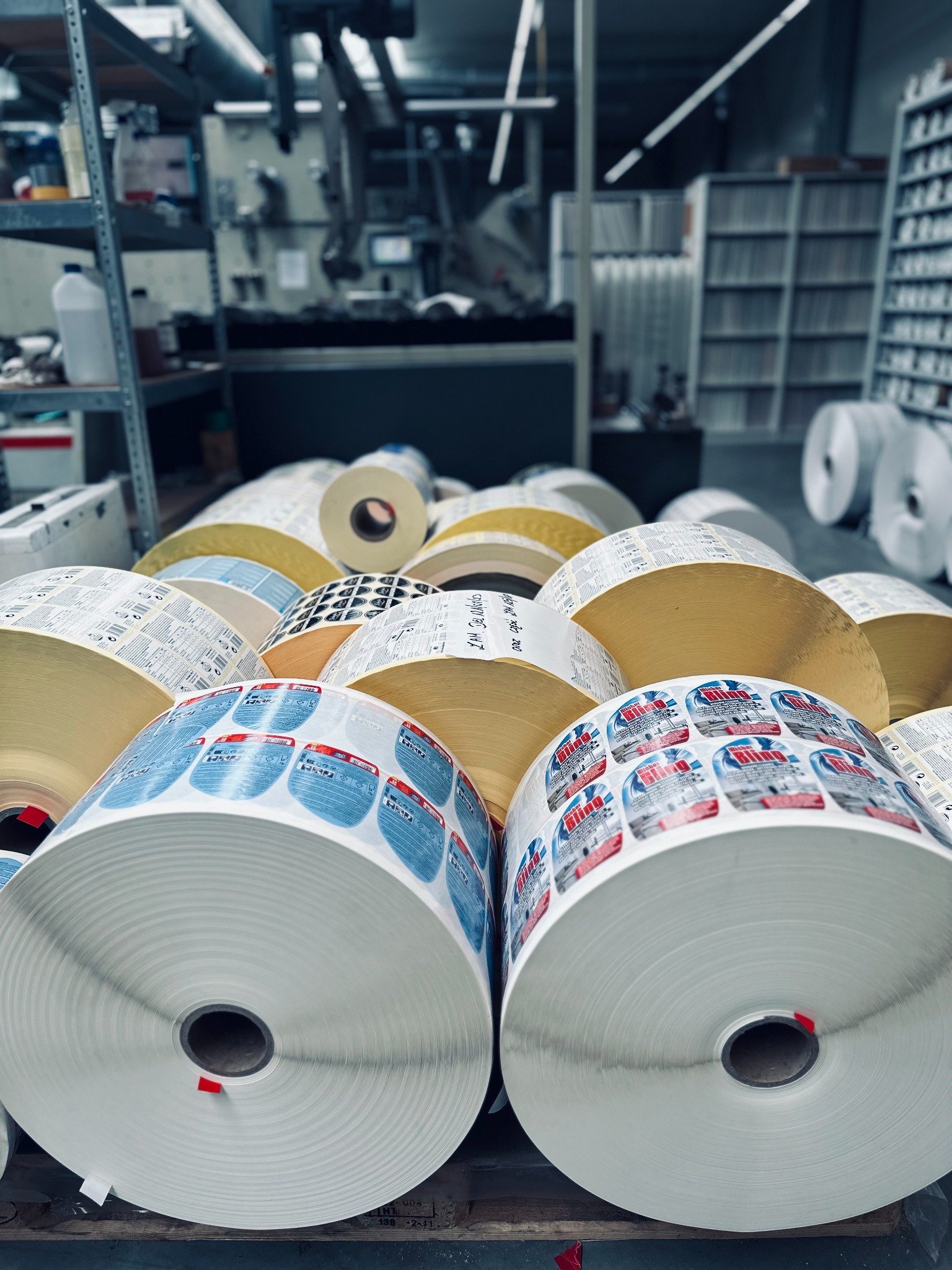Stos rolek z gotowymi etykietami w hali produkcyjnej drukarni, widoczne regały z materiałami eksploatacyjnymi w tle.