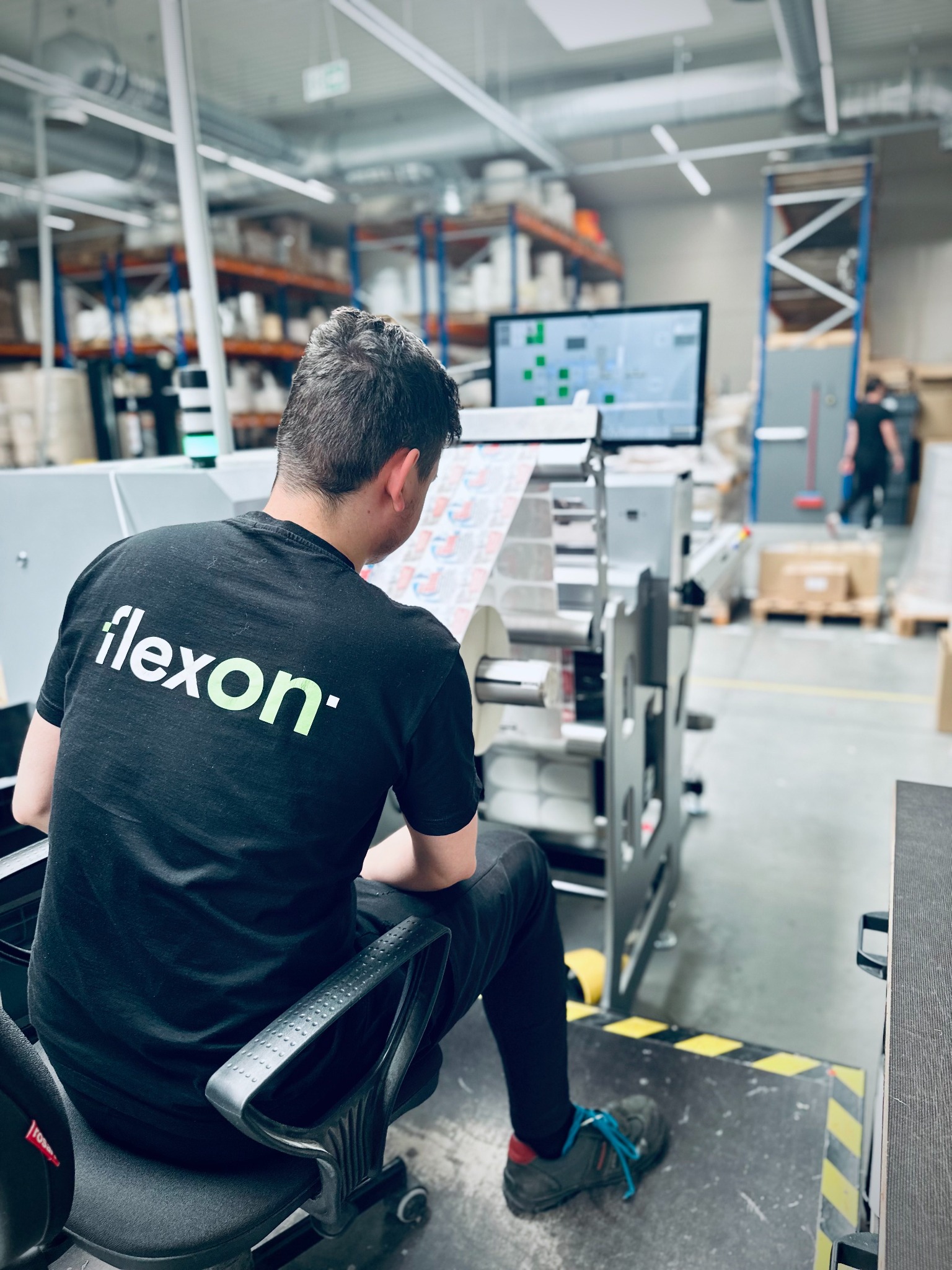 Pracownik firmy Flexon obsługuje maszynę drukarską, produkcja etykiet na roli, widoczne półki magazynowe w tle.