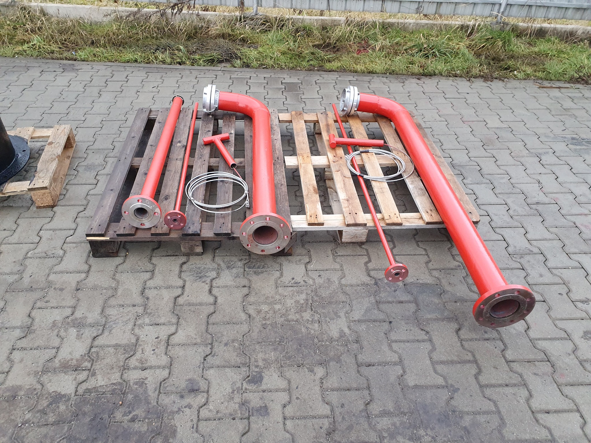 Czerwone rury z kołnierzami i narzędzia na drewnianej palecie, przygotowane do instalacji, widoczne na tle kostki brukowej.