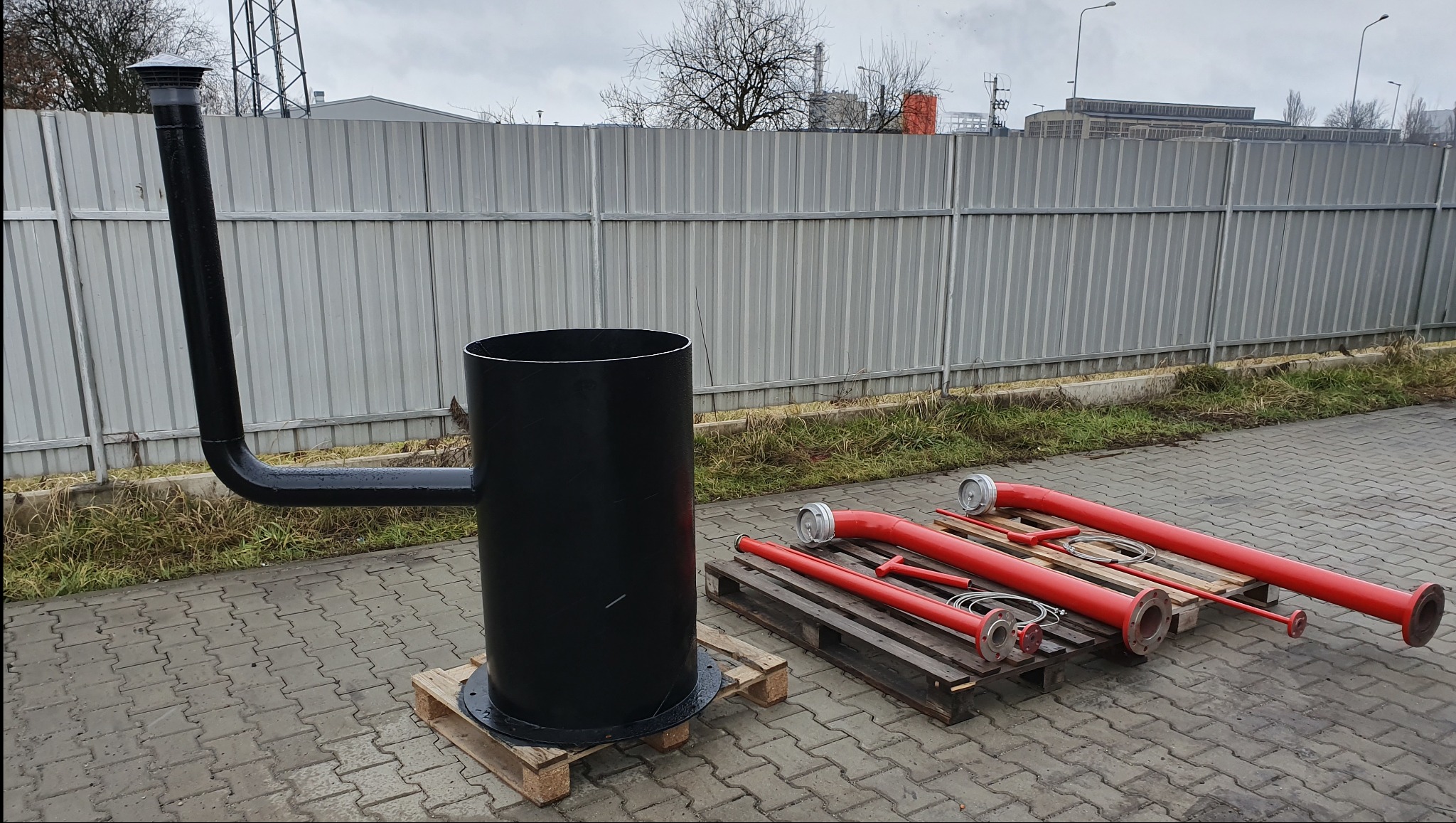 Czarny, metalowy zbiornik z rurą na palecie obok czerwonych rur strażackich na palecie, na zewnątrz, przy szarym metalowym ogrodzeniu.