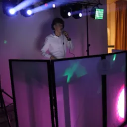 Młody DJ w białej koszuli, z mikrofonem i słuchawkami, za podświetlonym stanowiskiem DJ-skim, oświetlony reflektorami podczas występu.