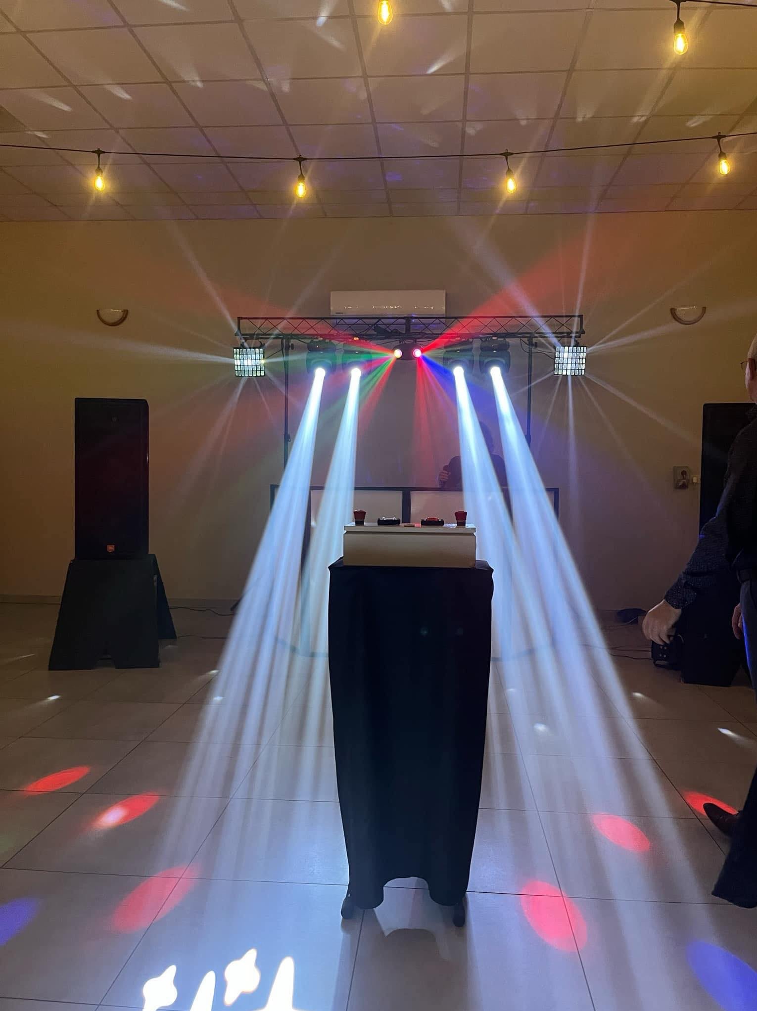 Sala bankietowa z profesjonalnym oświetleniem scenicznym i nagłośnieniem, widoczne reflektory, kolumny głośnikowe, stół DJ-a z kontrolerami, dekoracyjne żarówki na suficie i fragment sylwetki osoby...