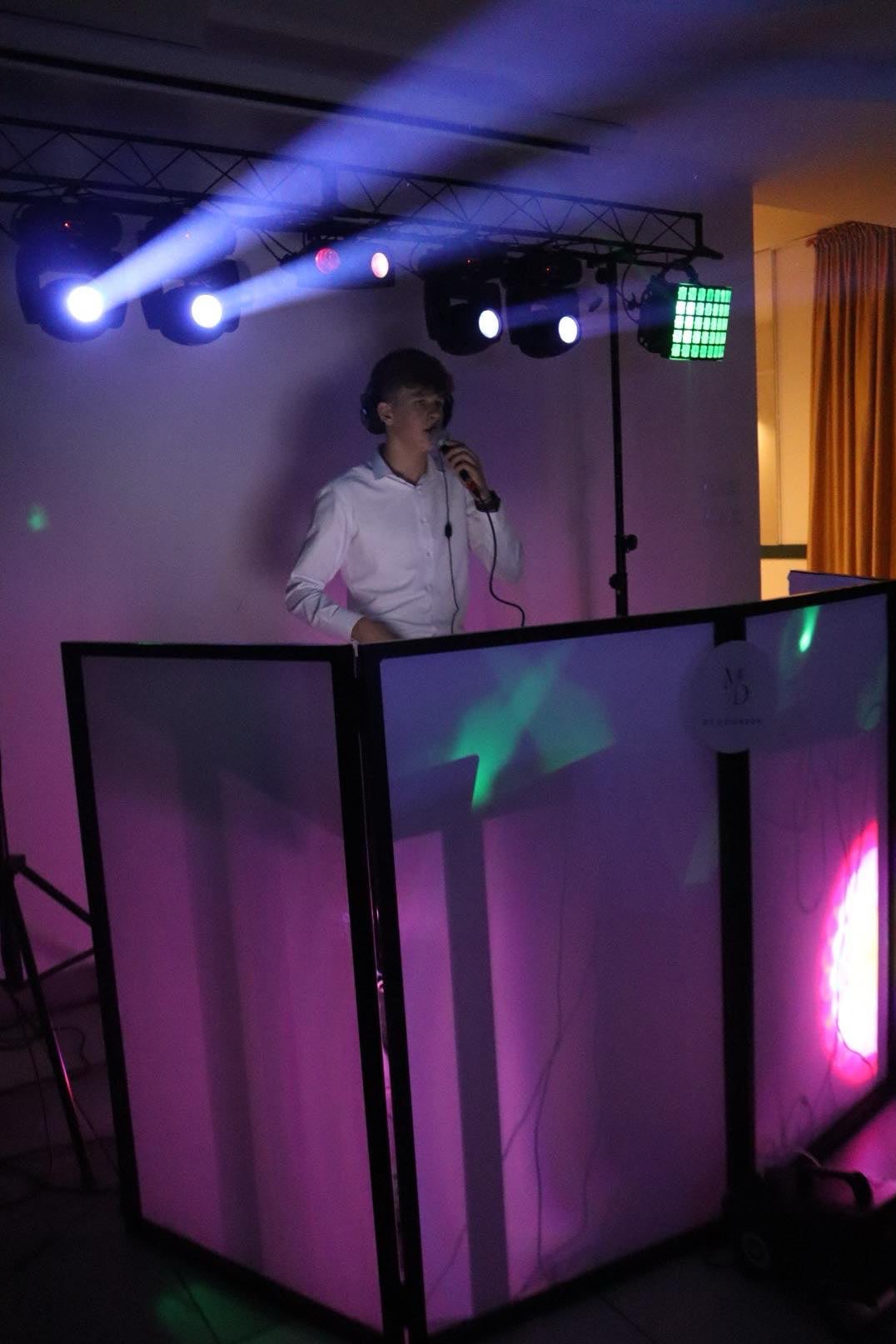 Młody DJ w białej koszuli, z mikrofonem i słuchawkami, za podświetlonym stanowiskiem DJ-skim, oświetlony reflektorami podczas występu.