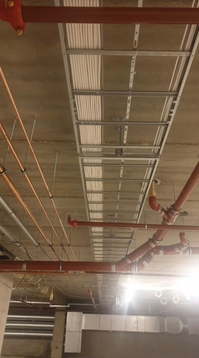 Sufit techniczny z metalowymi korytkami kablowymi wypełnionymi białymi przewodami, widoczne rury instalacji przeciwpożarowej i wentylacyjnej.