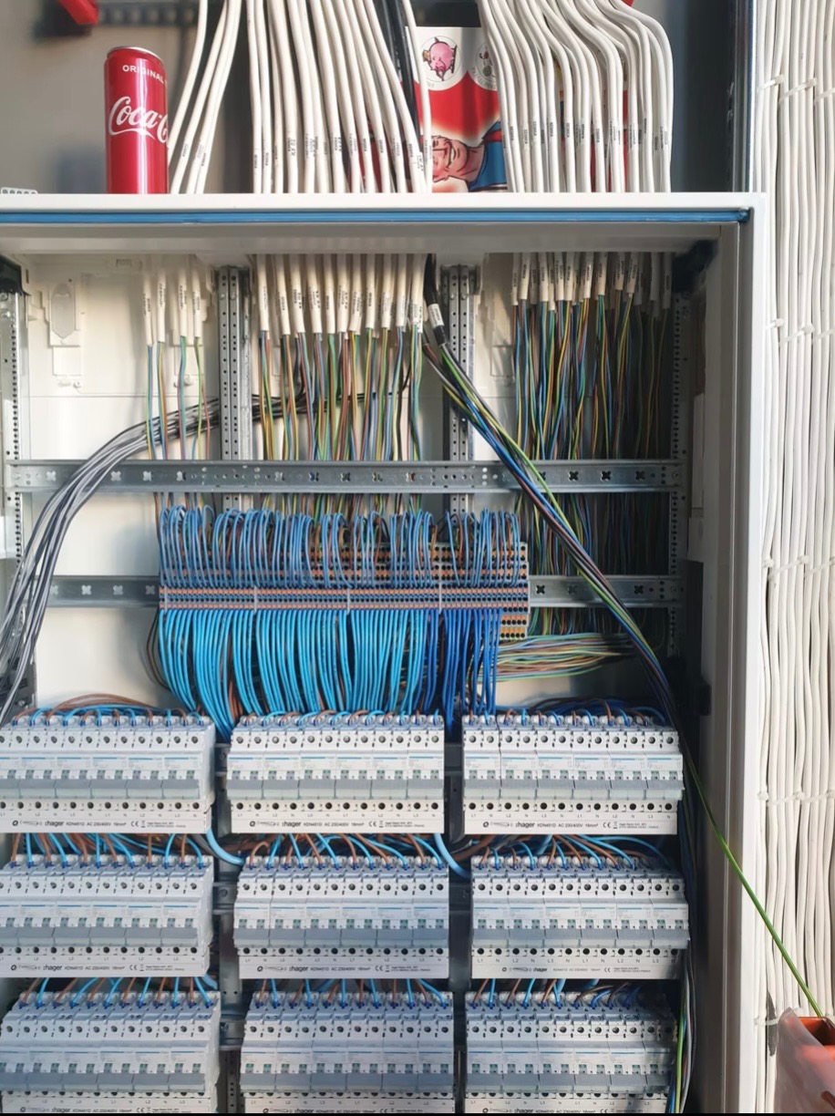 Rozdzielnica elektryczna z licznymi przewodami, zabezpieczeniami i listwami, na górze puszka Coca-Coli i pudełko po tabletkach.