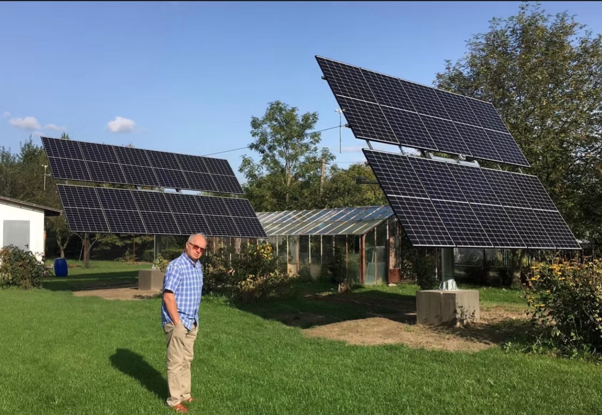 Dwa panele fotowoltaiczne ustawione na betonowych podstawach w ogrodzie, z widoczną szklarnią w tle i mężczyzną w okularach w pierwszym planie.