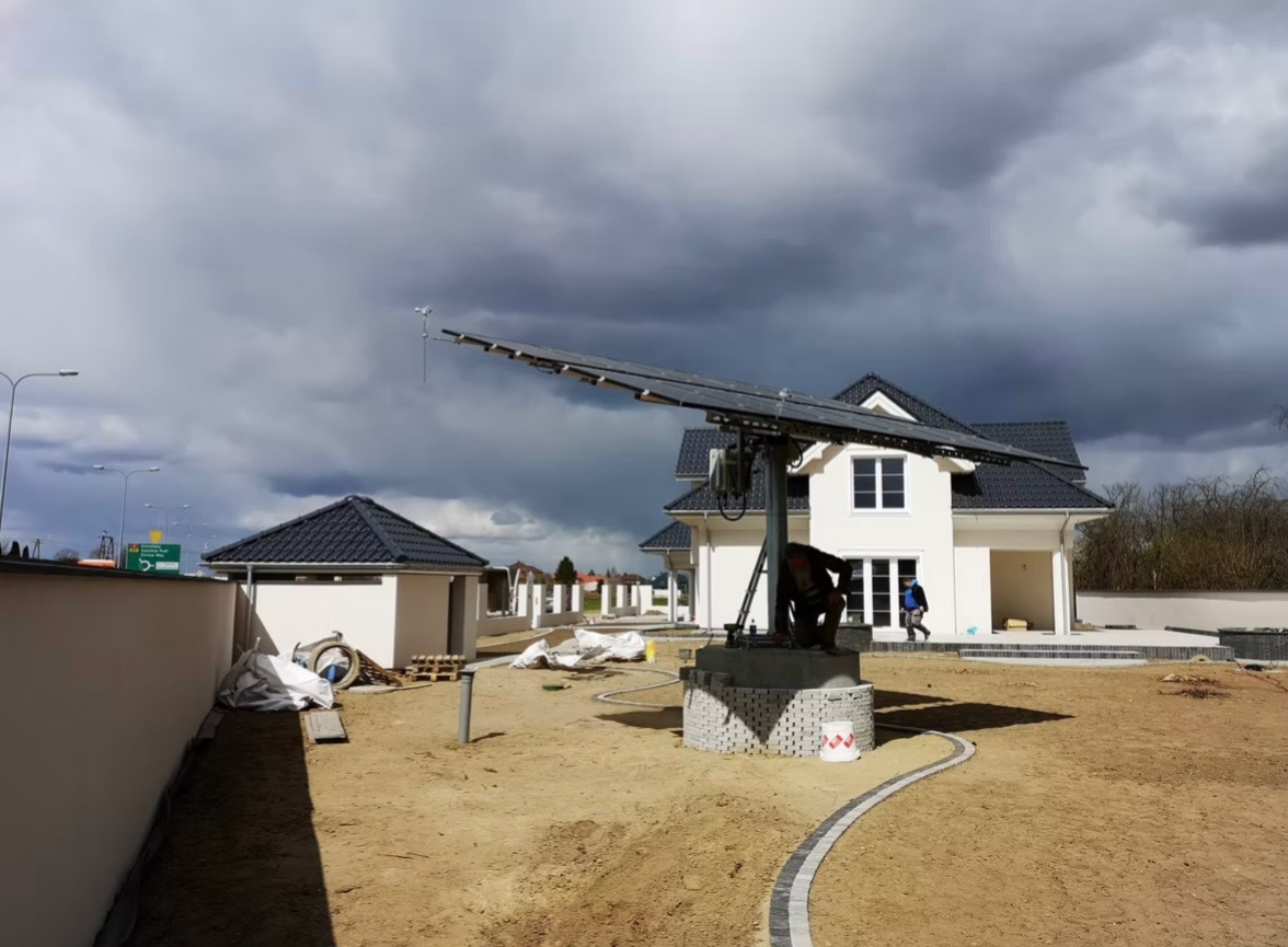 Montaż wysięgnika z panelami fotowoltaicznymi na posesji z nowym domem jednorodzinnym, widoczny pracownik przy konstrukcji i osoba wchodząca do domu.