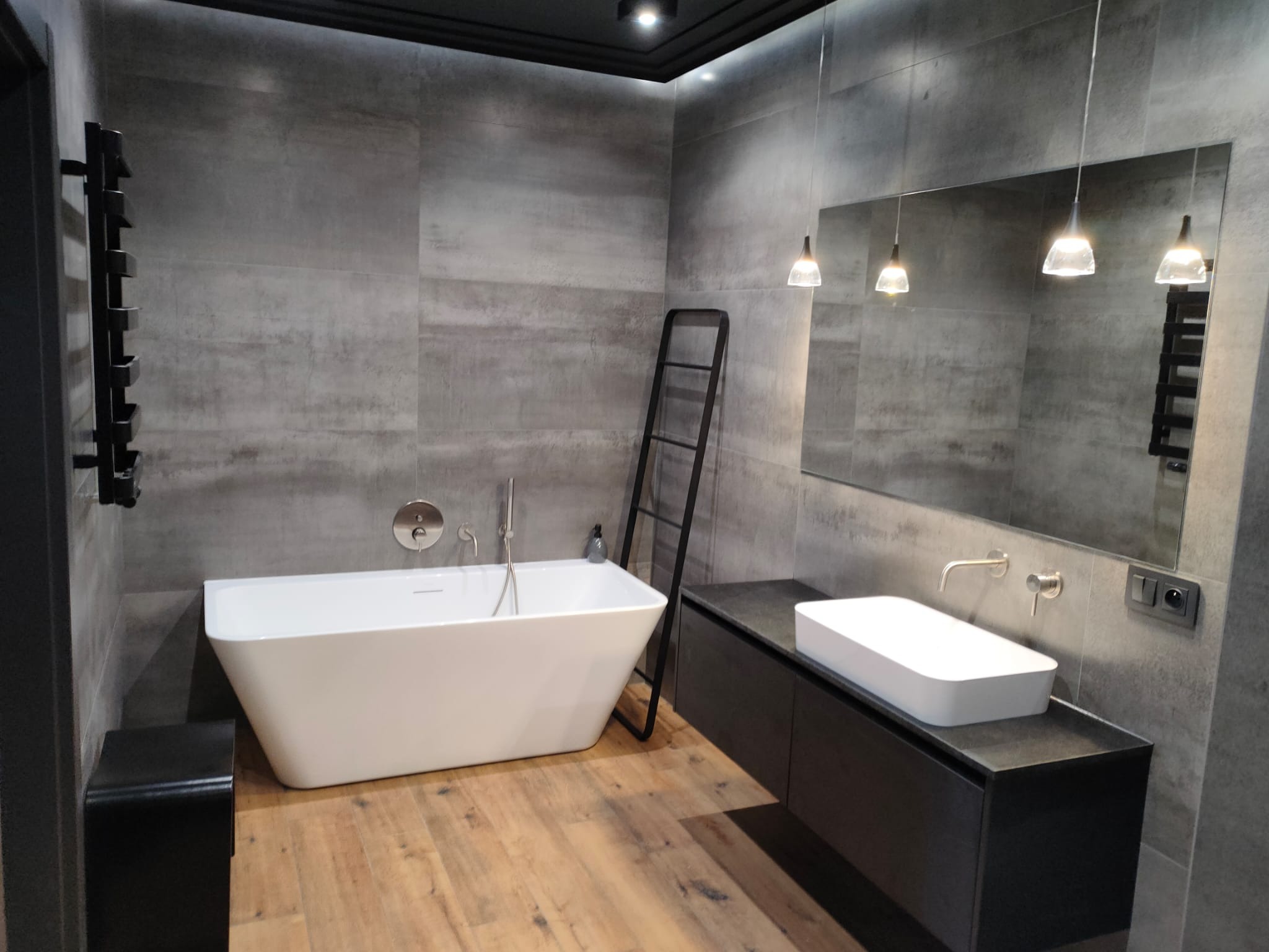 Nowoczesna łazienka z wanną wolnostojącą, szarą płytką imitującą beton, drewnianą podłogą i czarnymi akcentami, takimi jak drabinka na ręczniki i szafka pod umywalką.