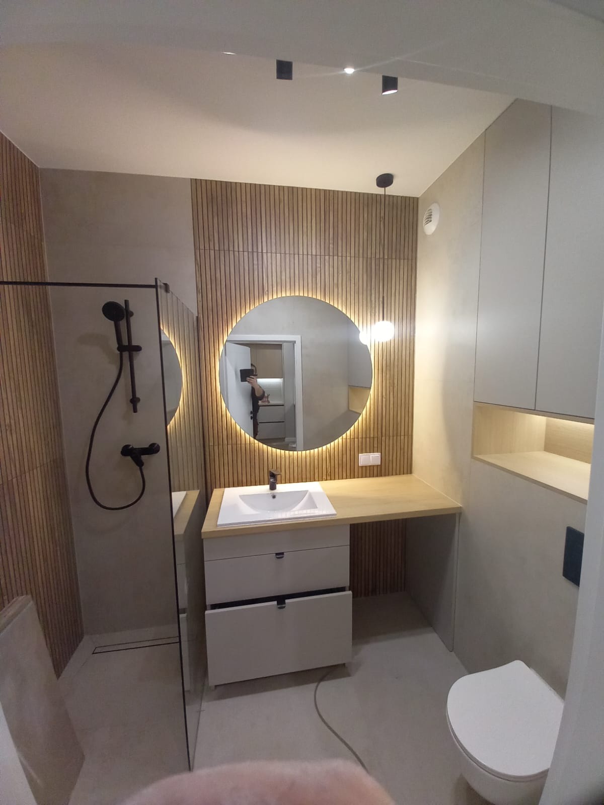 Nowoczesna łazienka z kabiną prysznicową typu walk-in, okrągłym lustrem z podświetleniem LED, umywalką nablatową z szafką i blatem z jasnego drewna oraz toaletą wiszącą.