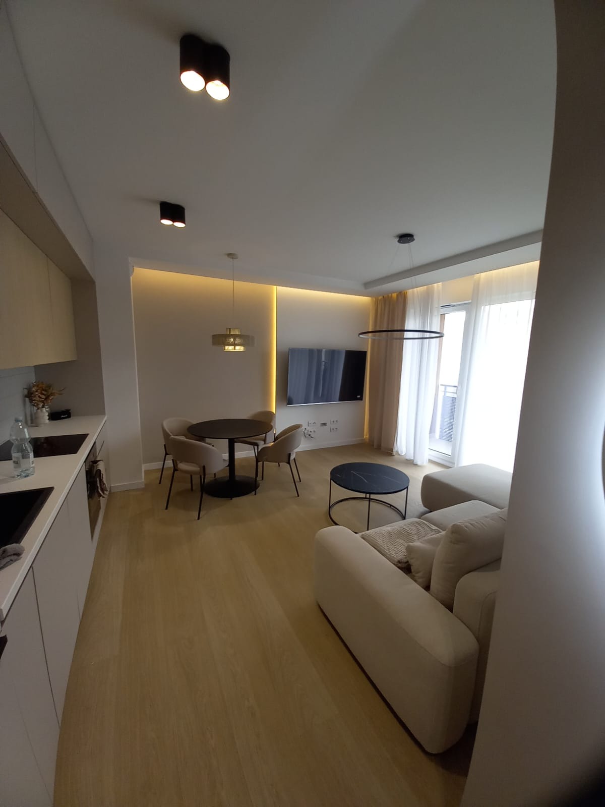 Jasne, przestronne wnętrze mieszkania z aneksem kuchennym, okrągłym stołem jadalnym i sofą, oświetlone lampami sufitowymi i taśmami LED.