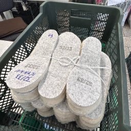 P.P.H.U. Jolanta Leśniak - Stosy szarych filcowych wkładek do obuwia w zielonym plastikowym pojemniku, z widocznymi rozmiarami naniesionymi markerem, w tle widoczny stół i maszyna do szycia.