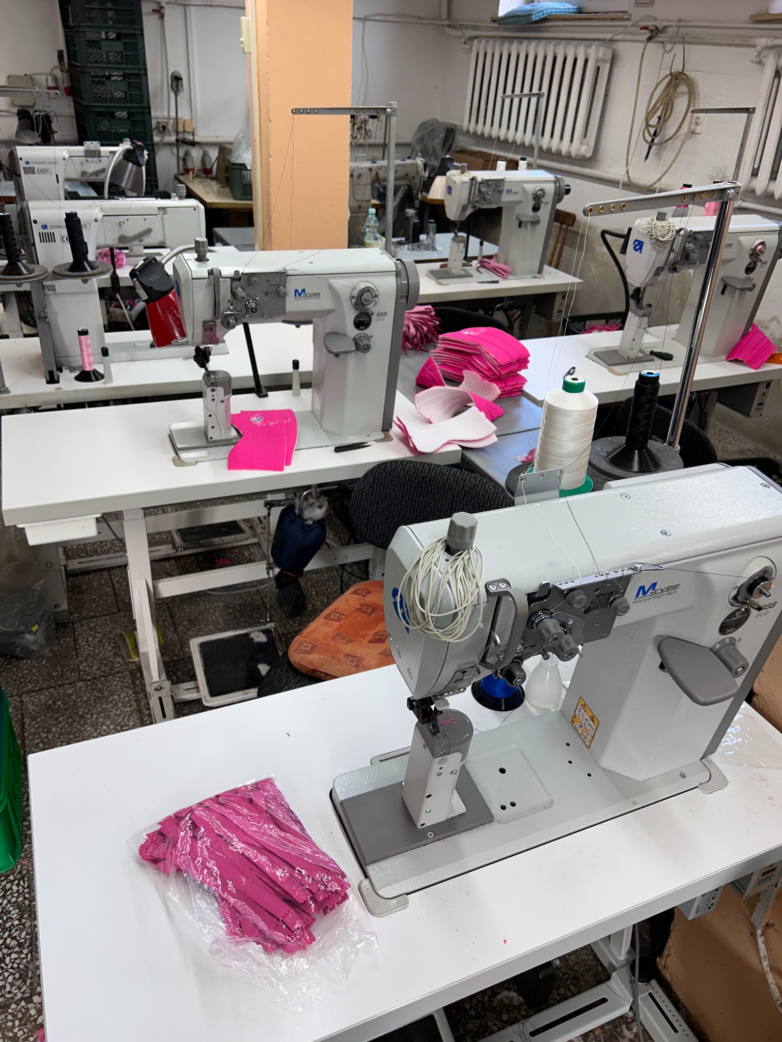 Wnętrze szwalni z rzędem maszyn do szycia, różowe elementy materiałów i nici na stołach roboczych, widoczne detale wyposażenia i oświetlenia.