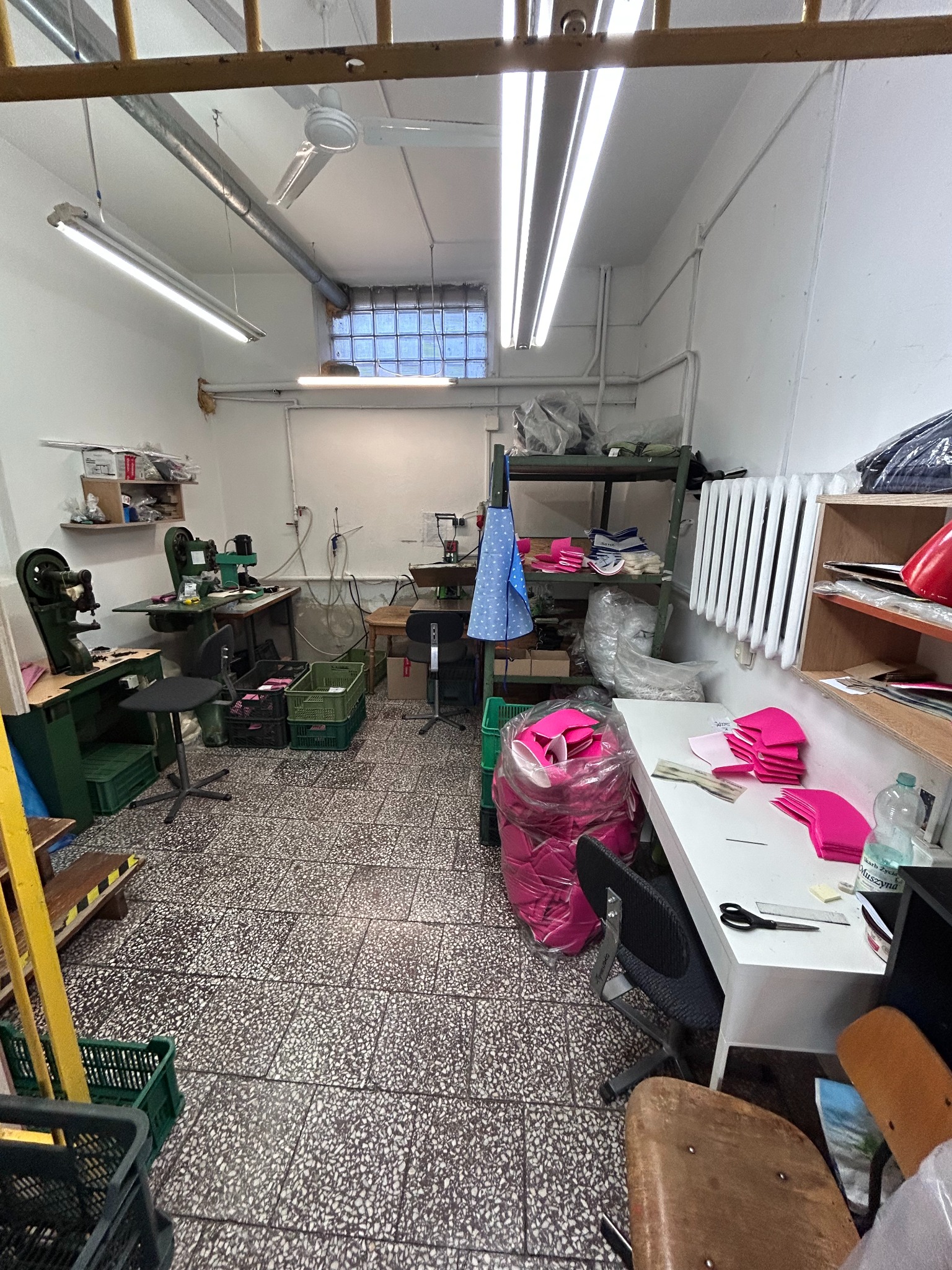 Wnętrze szwalni z maszynami do szycia, stołami roboczymi z rozłożonymi elementami z różowej tkaniny, prawdopodobnie do szycia pokrowców lub pościeli, widoczne także kosze z materiałami i regały...