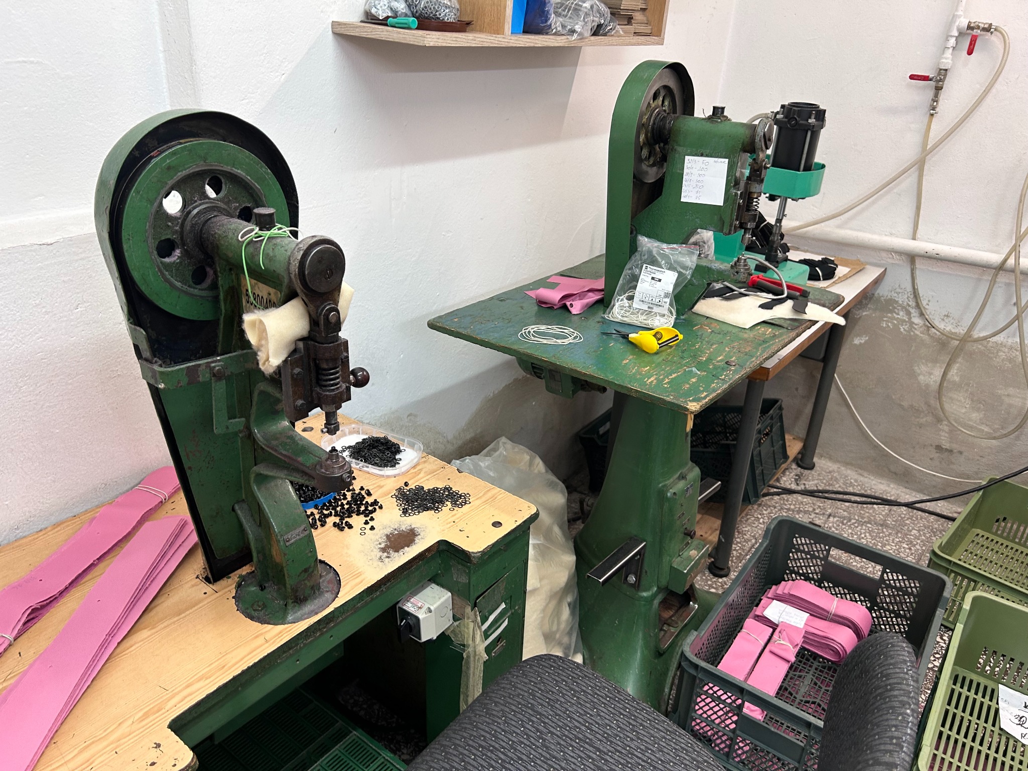 Dwie stare, zielone maszyny szwalnicze w warsztacie. Jedna maszyna z czarnymi nitami i różowymi paskami materiału, druga z nożyczkami i gumkami na stole. Widoczne plastikowe pojemniki z materiałami...