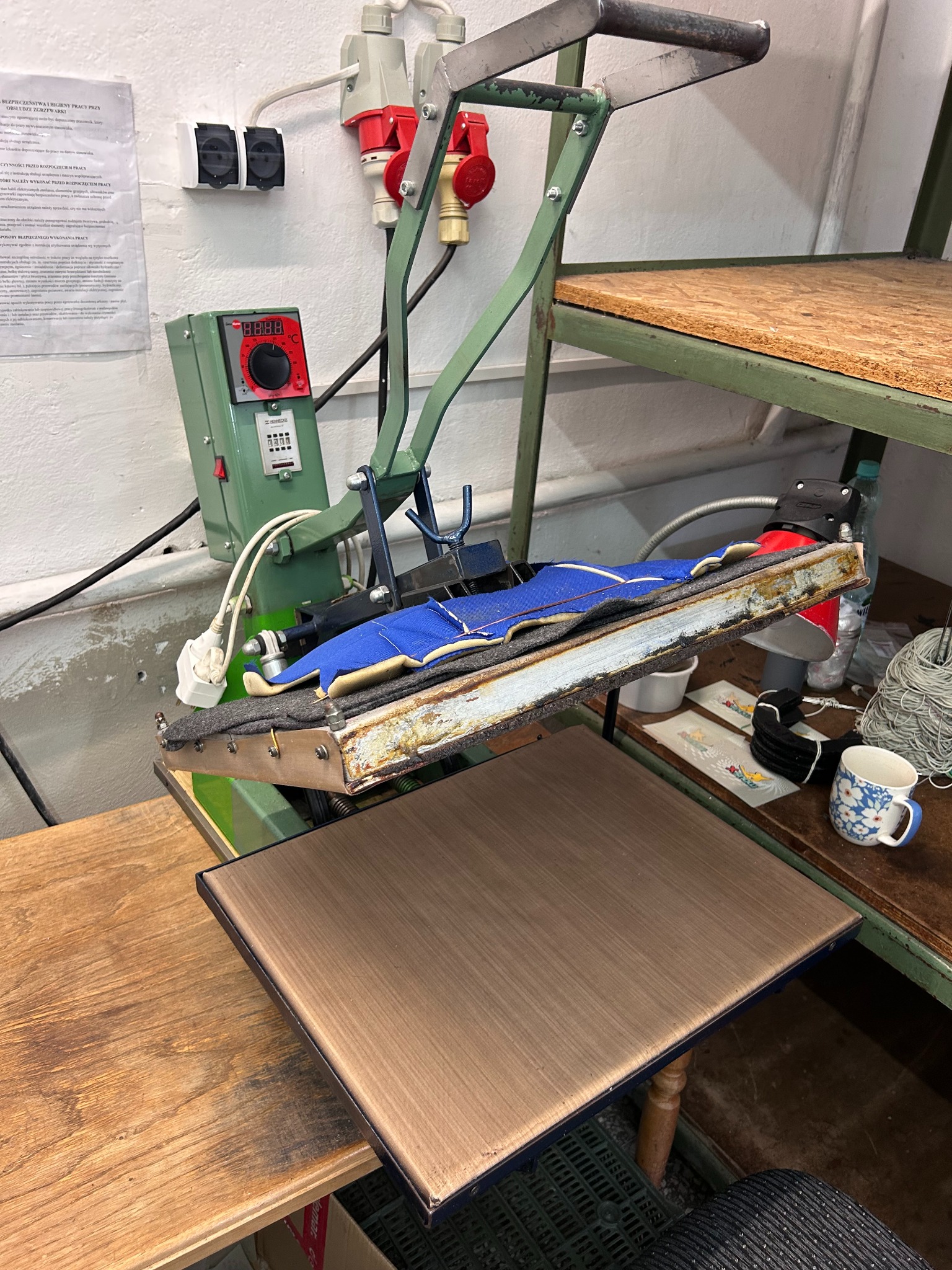 Przemysłowa prasa termotransferowa z niebieskim elementem odzieży roboczej, widoczne przewody, panel sterowania i otoczenie warsztatowe.