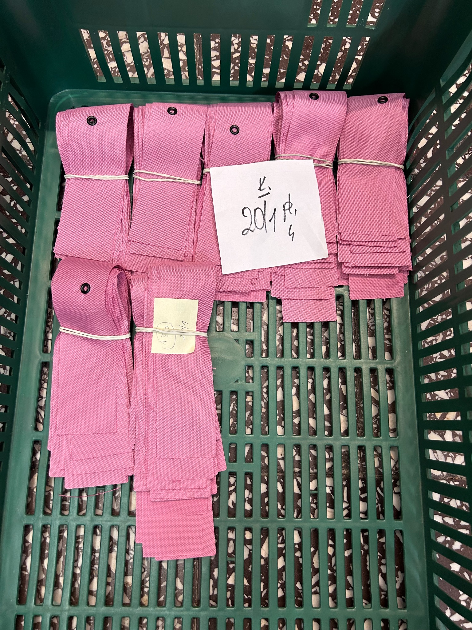 Różowe paski materiału z oczkiem, spięte gumką, ułożone w zielonym koszu. Dołączone karteczki z odręcznymi notatkami.