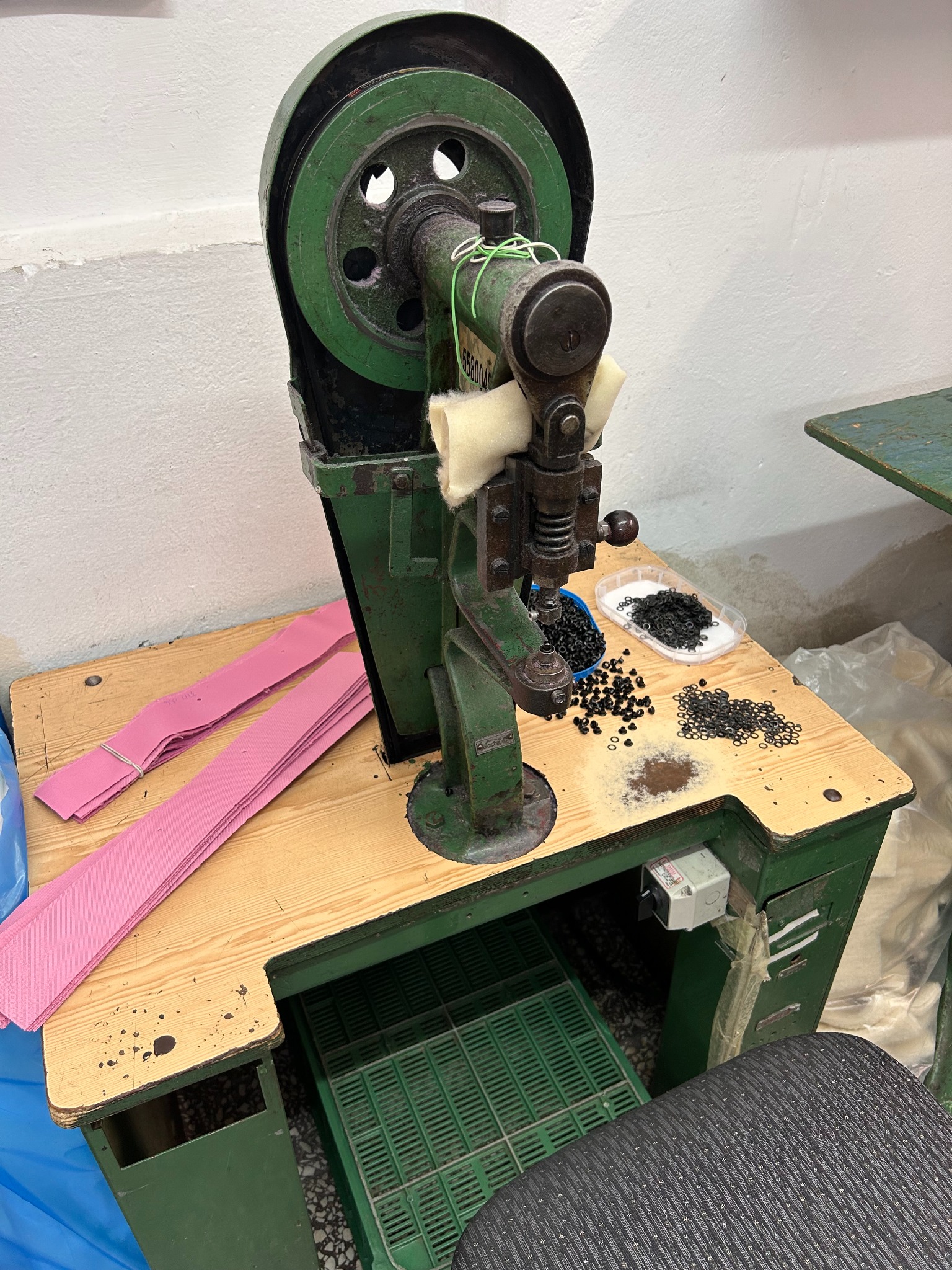 Starodawna, zielona maszyna do nabijania nap na stole warsztatowym z rozrzuconymi czarnymi napami i stosem różowych pasków materiału.