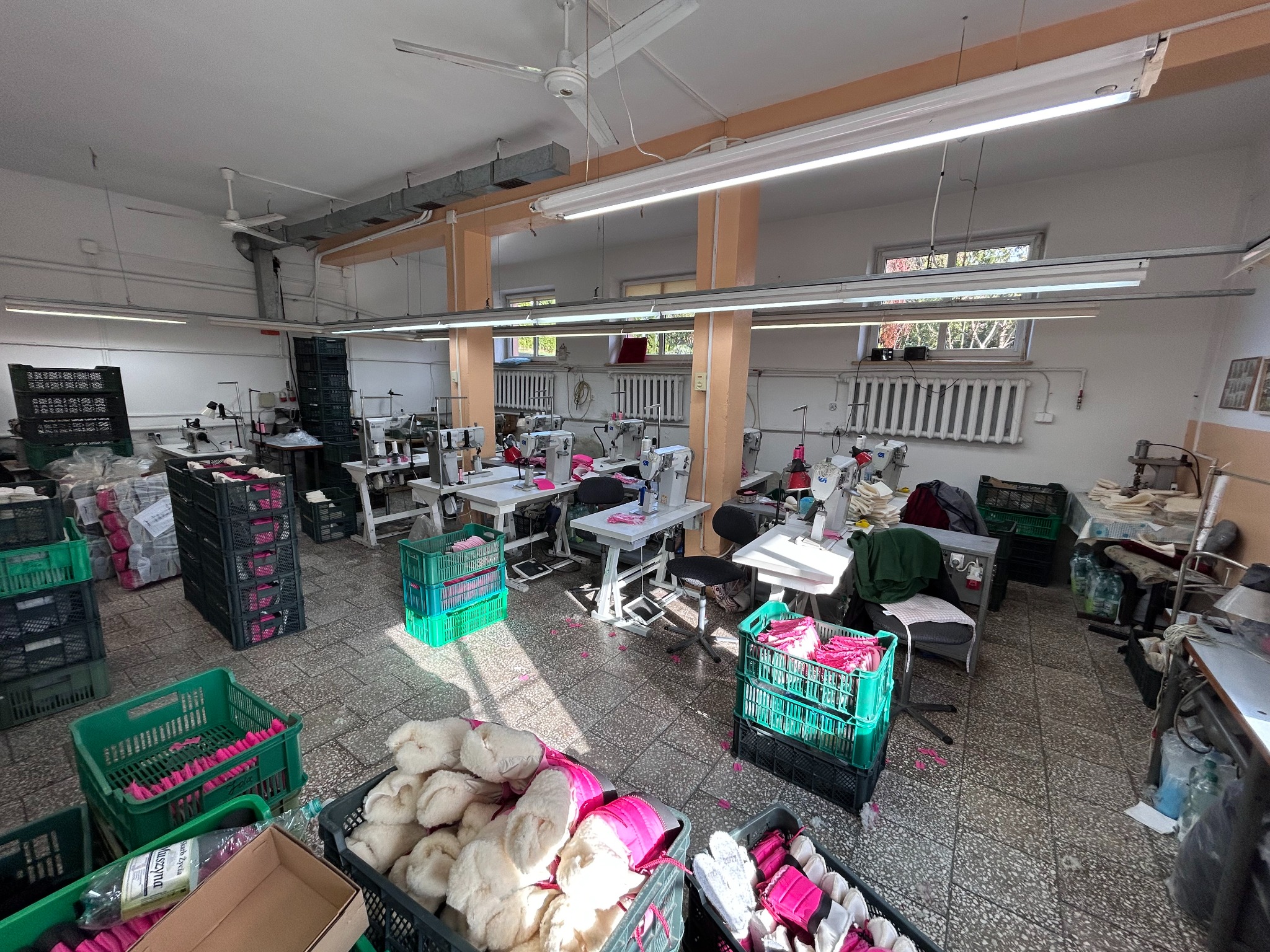 Widok hali produkcyjnej z licznymi maszynami do szycia, plastikowymi pojemnikami z elementami odzieży i gotowymi produktami, oświetlonej świetlówkami.