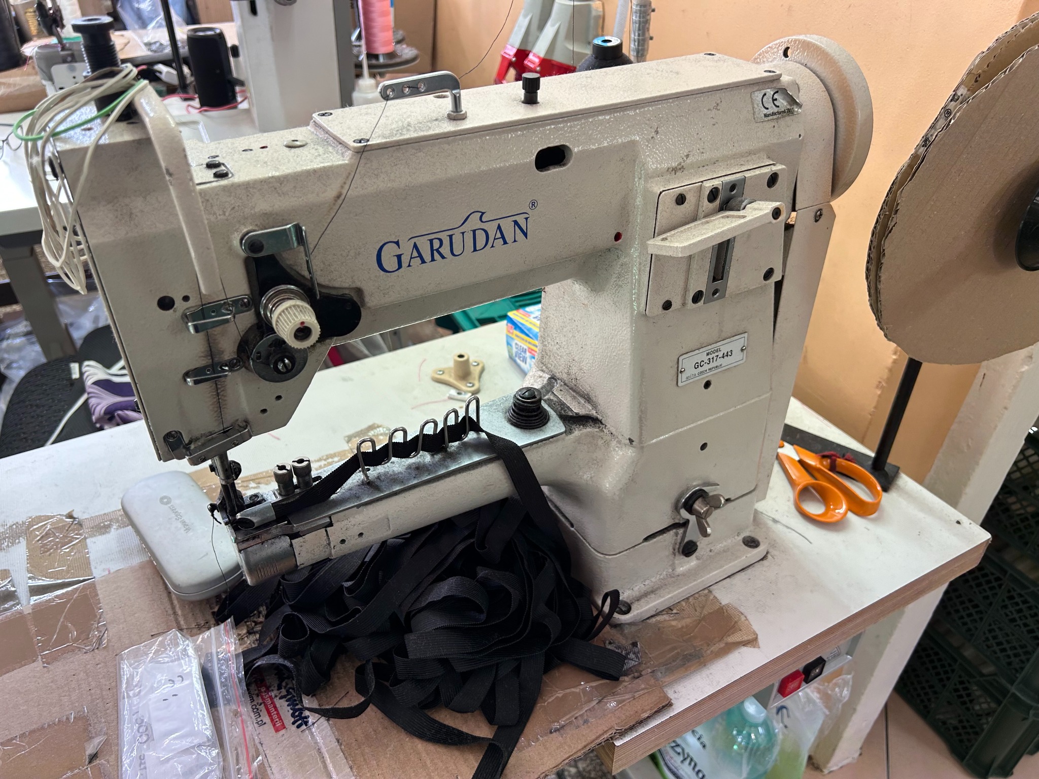Przemysłowa maszyna do szycia marki Garudan, model GC-317-443, z czarną taśmą pod igłą, nożyczkami i akcesoriami na stole roboczym.