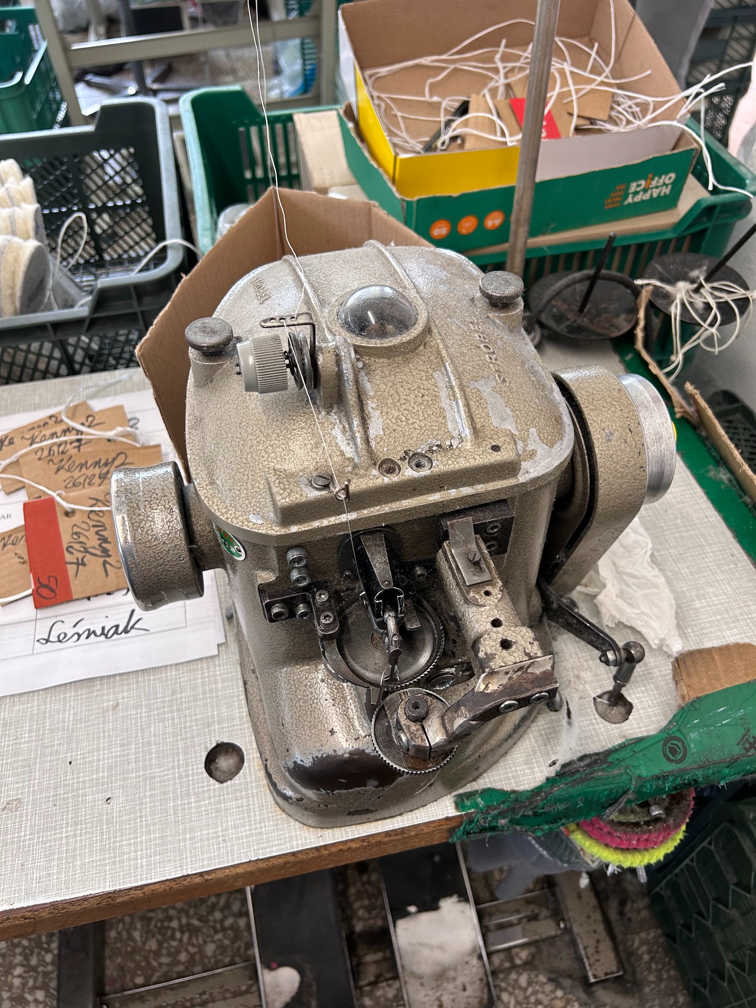 Starsza, używana maszyna szwalnicza, prawdopodobnie szewska, z widocznymi śladami użytkowania, w otoczeniu materiałów i narzędzi szwalniczych, na stole roboczym z widocznymi plamami i zabrudzeniami.