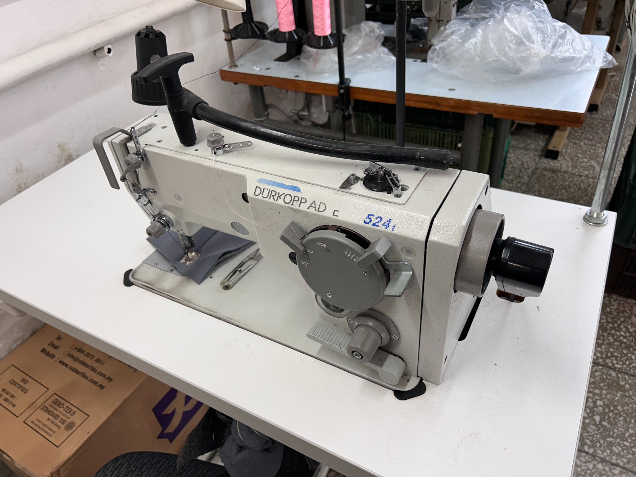 Profesjonalna maszyna do szycia Dürkopp Adler 524i na białym stole, z widocznymi detalami mechanizmu i fragmentem szarego materiału pod stopką.