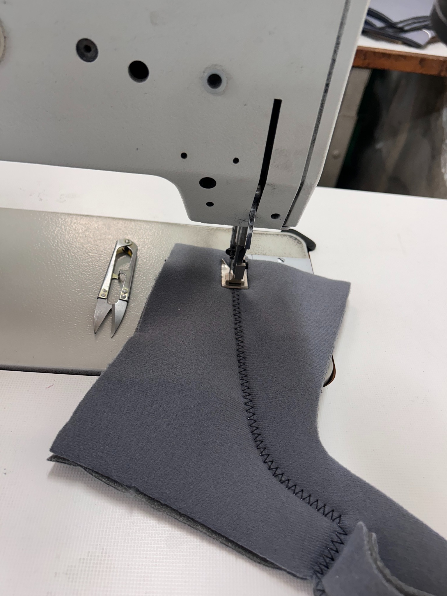 Fragment szarego materiału pod igłą maszyny do szycia, widoczne ściegi i nożyczki do nici po lewej stronie.