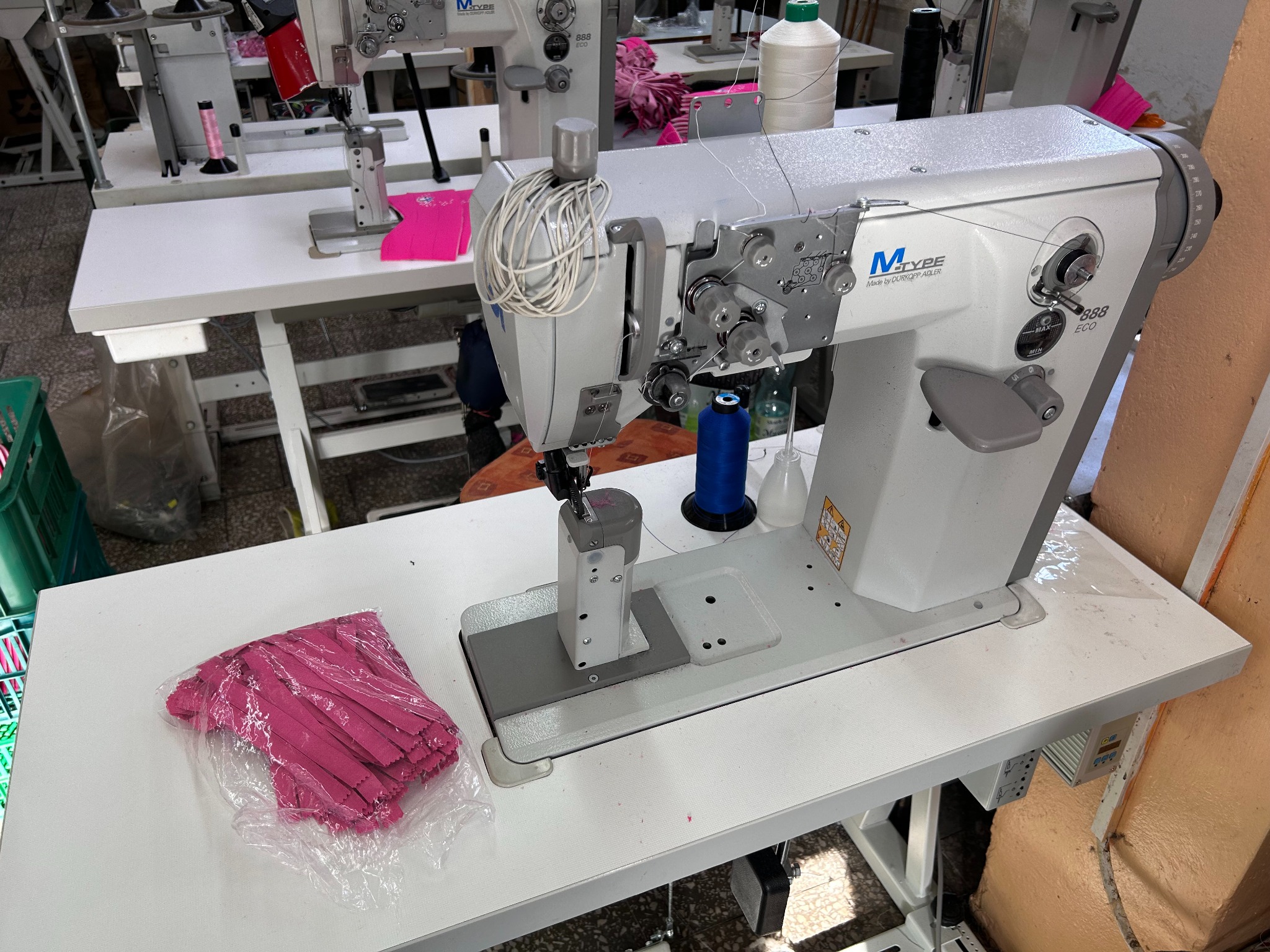 Przemysłowa maszyna do szycia M-Type 888 ECO z niebieską nicią i stosem różowych elementów materiału w przezroczystej folii na białym stole, w tle inne maszyny i akcesoria szwalnicze.