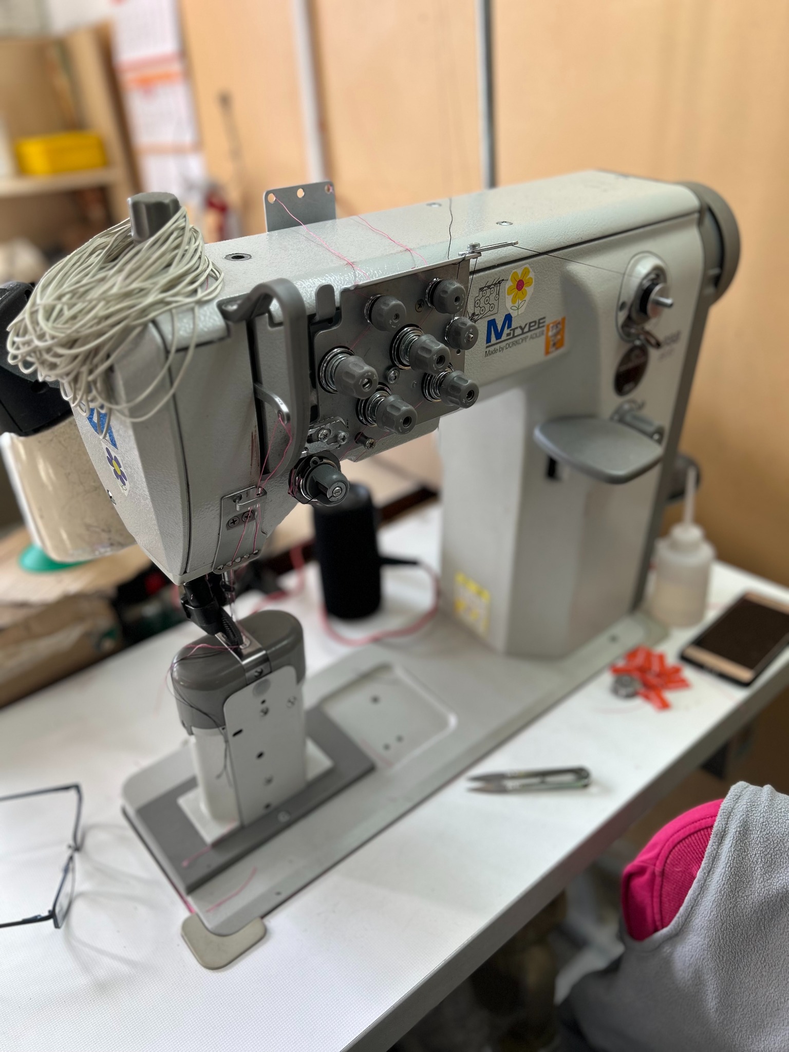 Profesjonalna maszyna do szycia M-TYPE z nawiniętą nicią, nożyczkami, telefonem i okularami na stole roboczym w szwalni.