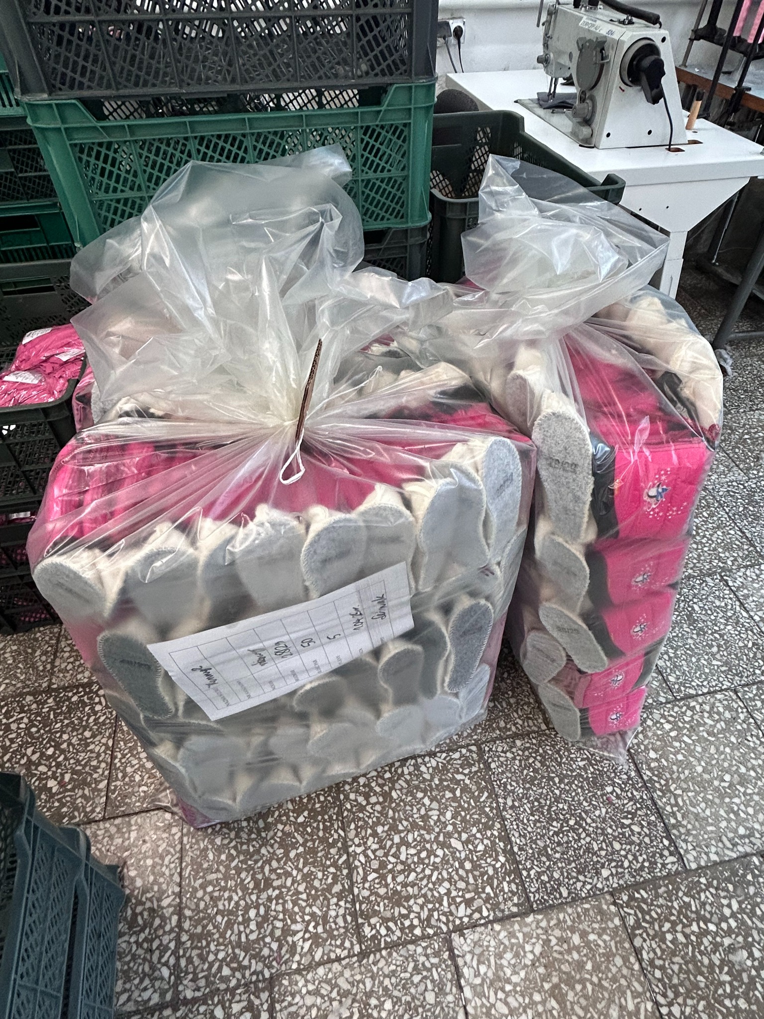 Dwa worki foliowe wypełnione gotowymi, uszytymi kapciami dziecięcymi z różowym wykończeniem i szarą podeszwą, jeden worek z etykietą, w tle maszyna do szycia i plastikowe skrzynki.