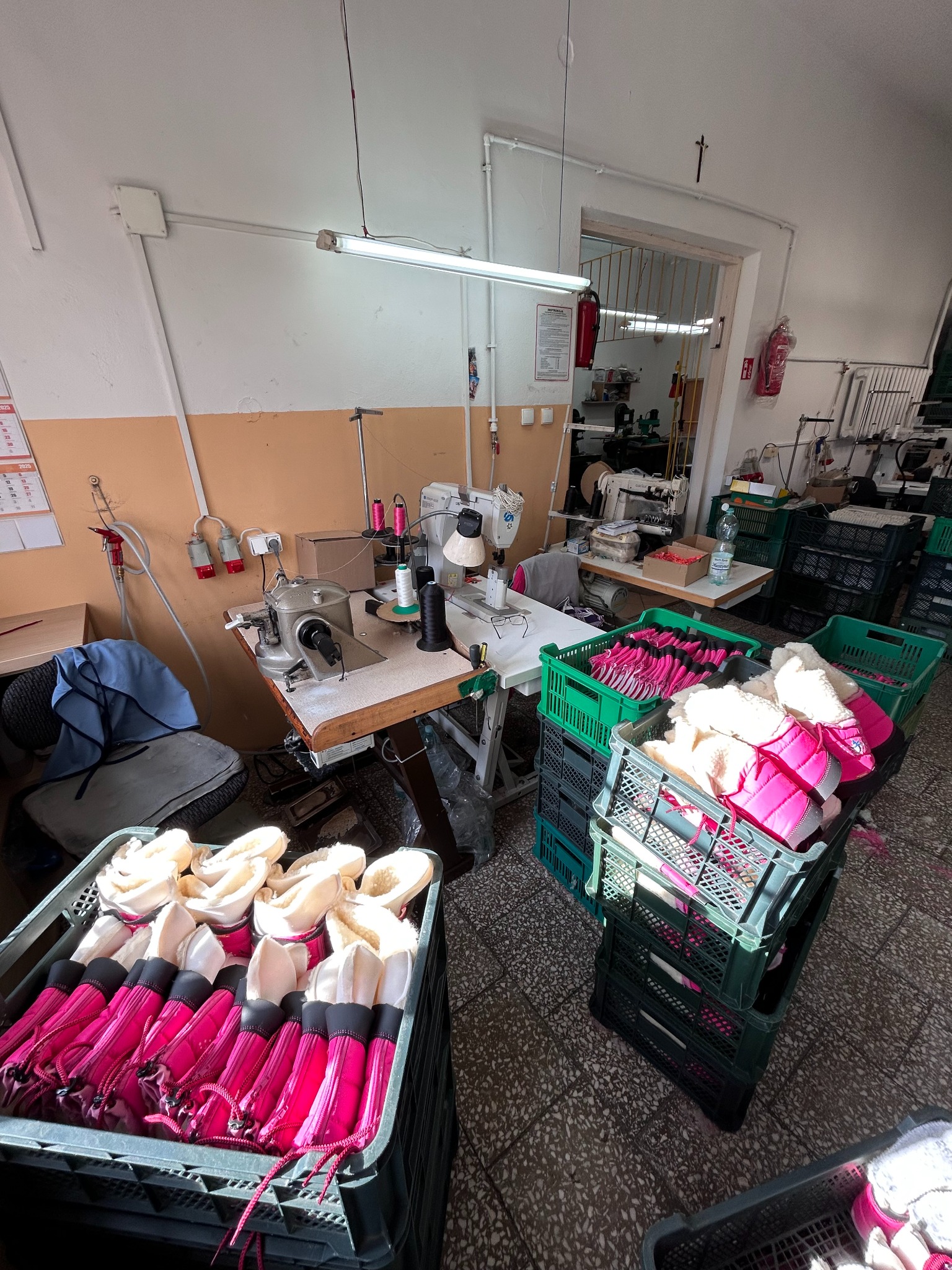 Wnętrze szwalni z maszynami do szycia i skrzynkami wypełnionymi gotowymi ocieplanymi pokrowcami na buty z różowymi elementami, widok z góry.