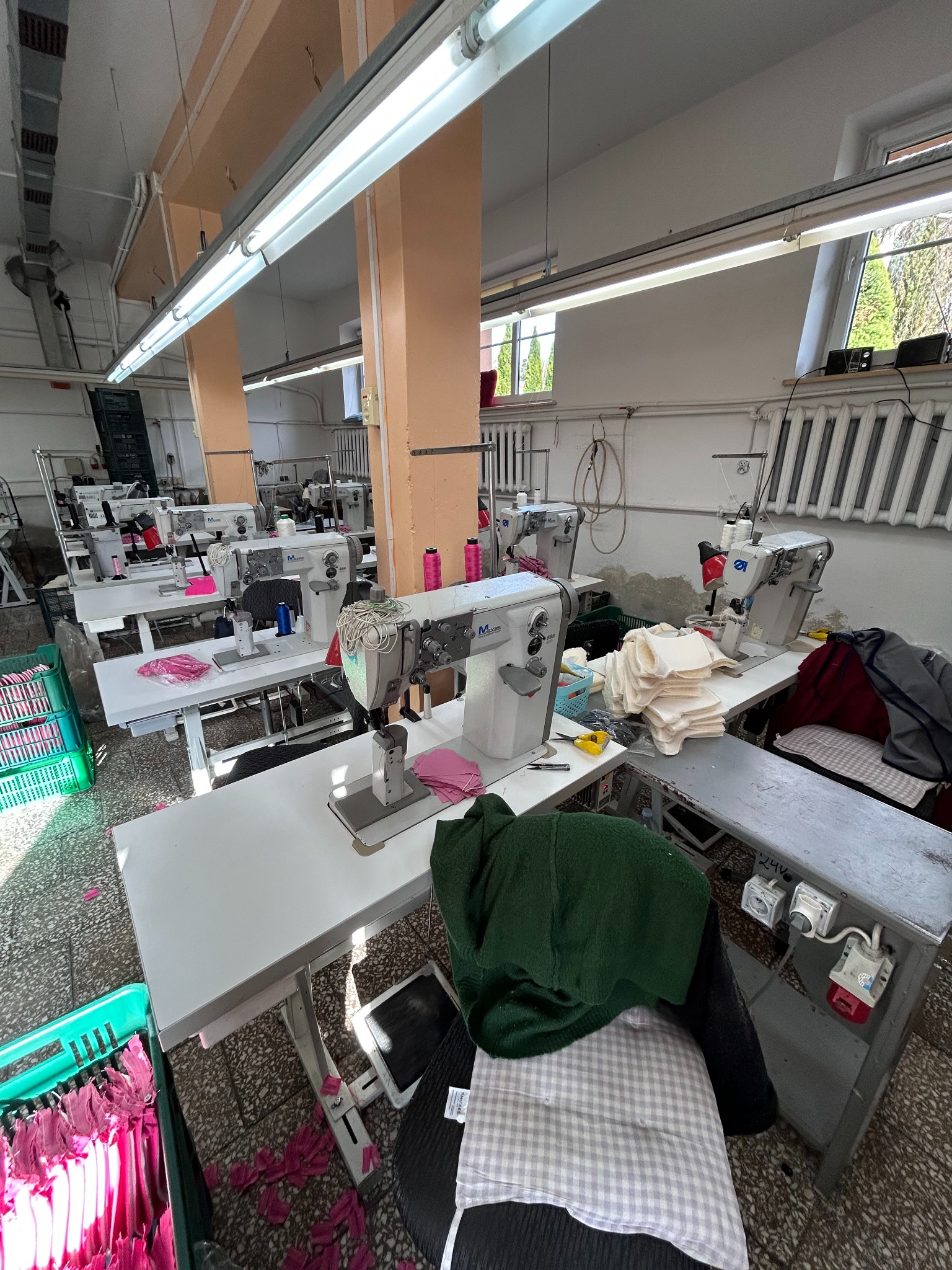 Wnętrze szwalni z rzędami maszyn do szycia, kosze z różowymi elementami, widoczne okna z kratami i instalacje wentylacyjne pod sufitem.