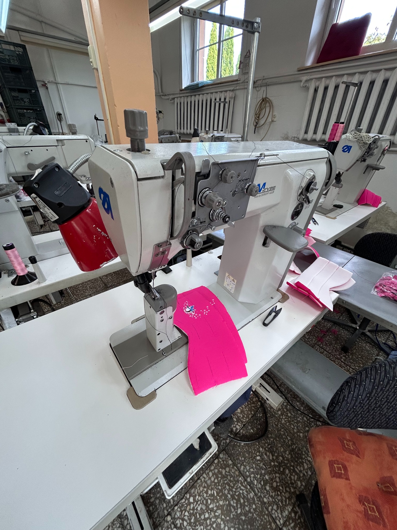 Szwalnia: Przemysłowa maszyna do szycia z czerwonym dodatkowym oświetleniem, szyjąca element z różowego materiału z nadrukiem gwiazdek, na tle inne maszyny i okno.