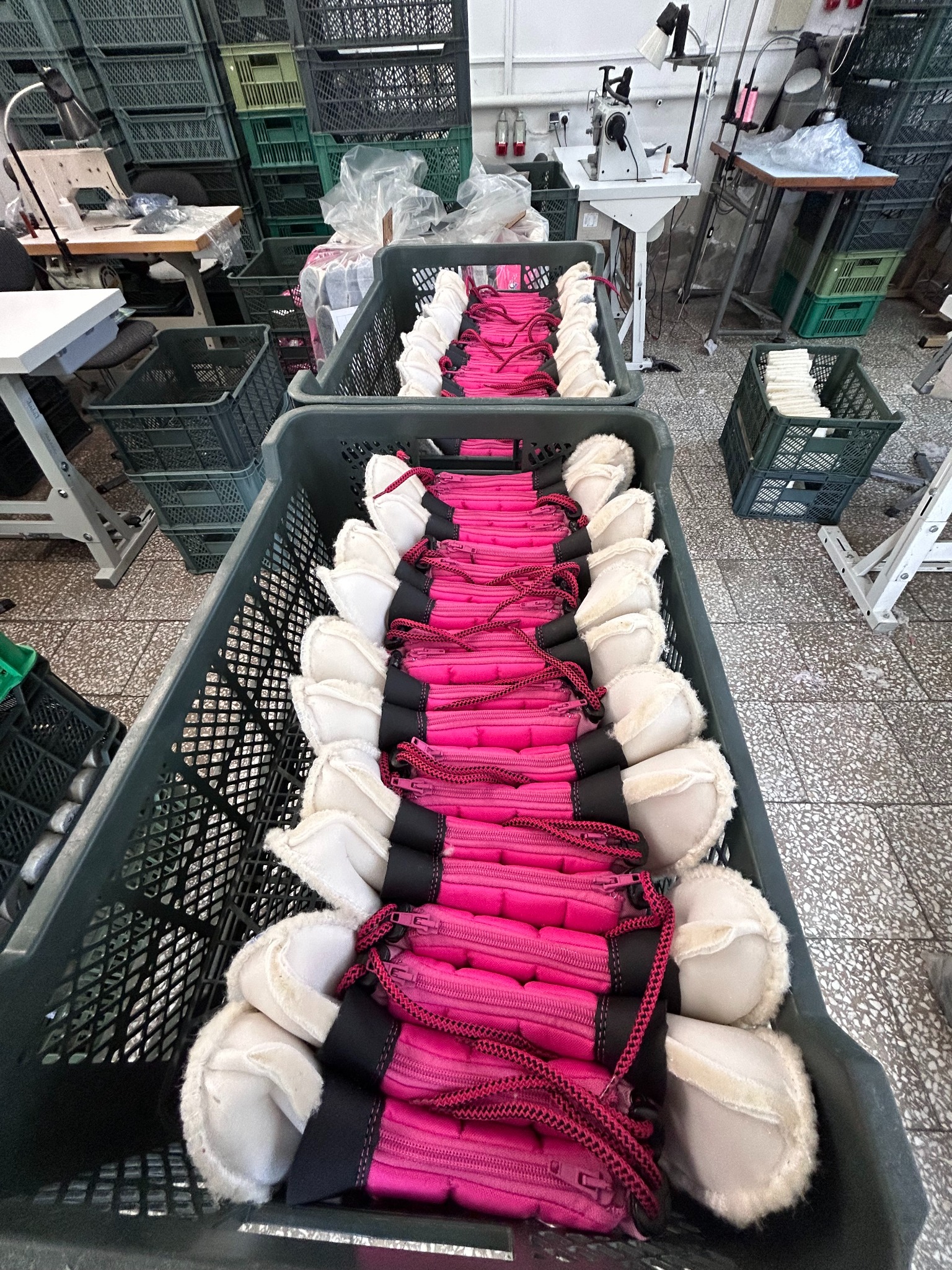 Rząd różowych pokrowców z białymi, futerkowymi wstawkami, ułożonych w plastikowych skrzynkach w szwalni, widoczne maszyny do szycia i stos skrzynek w tle.