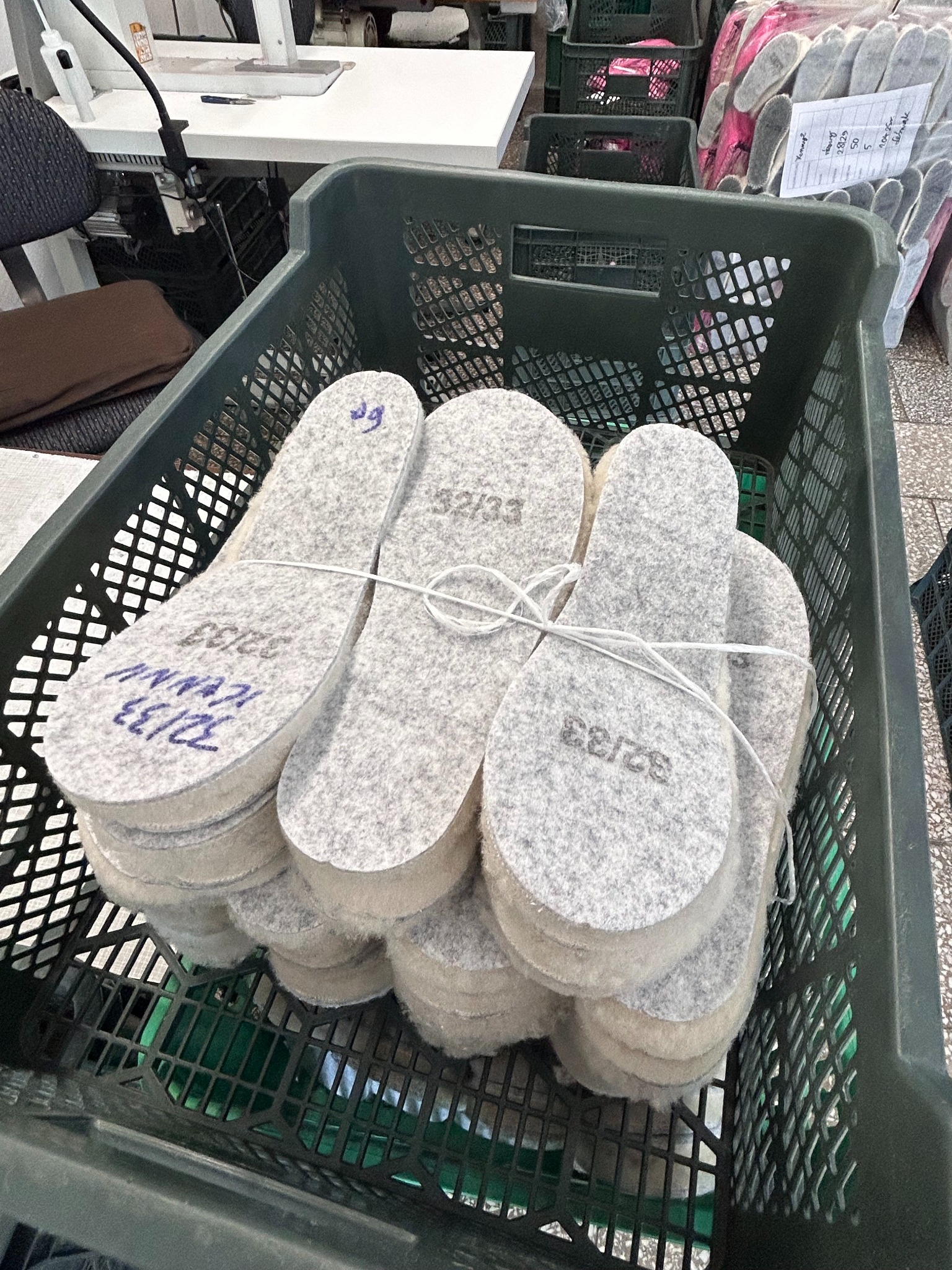 Stosy szarych filcowych wkładek do obuwia w zielonym plastikowym pojemniku, z widocznymi rozmiarami naniesionymi markerem, w tle widoczny stół i maszyna do szycia.