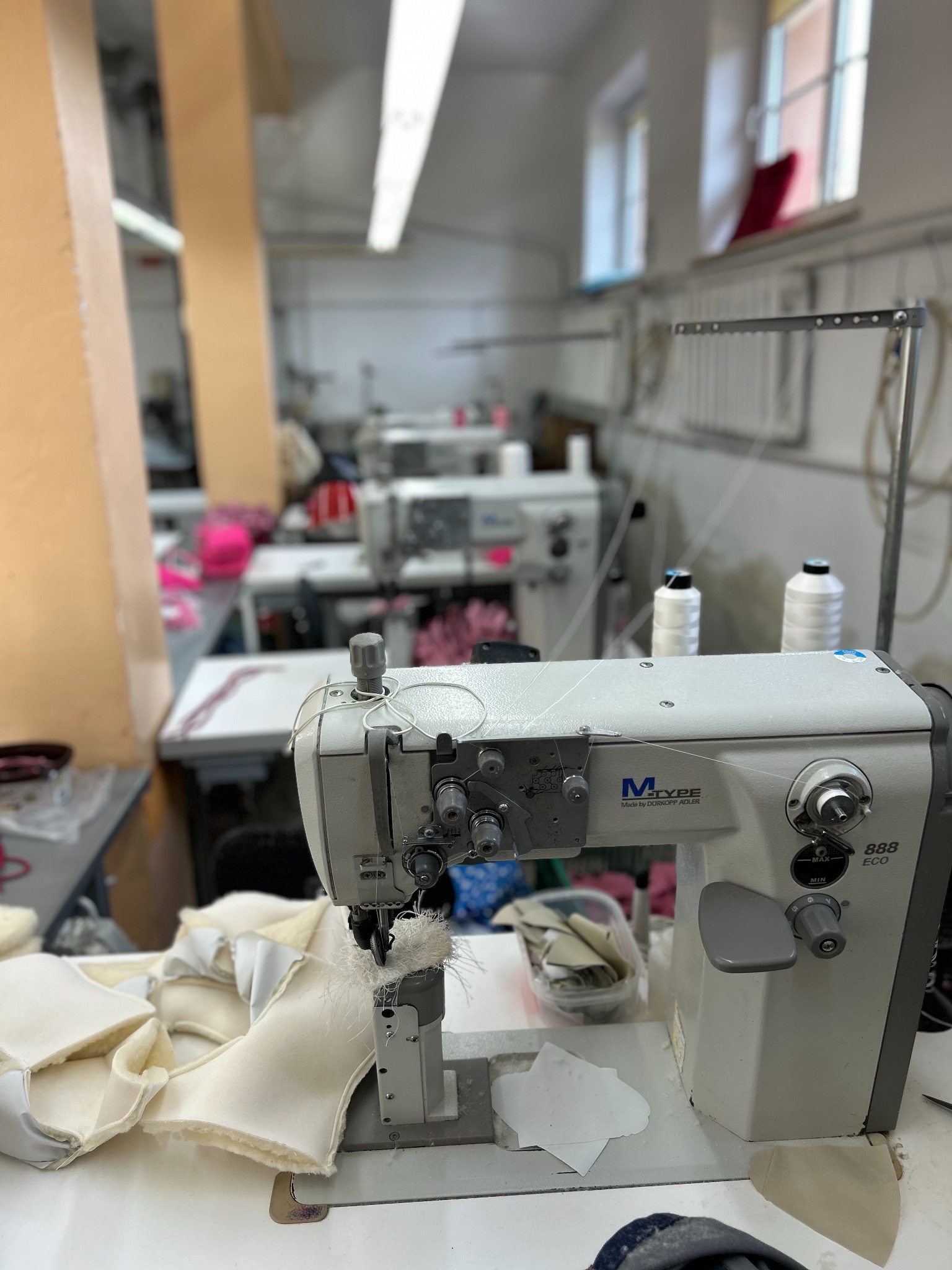 Szwalnia: Maszyna do szycia M-Type Durkopp Adler 888 ECO w trakcie szycia jasnego materiału z futerkowym wykończeniem, w tle inne stanowiska pracy.
